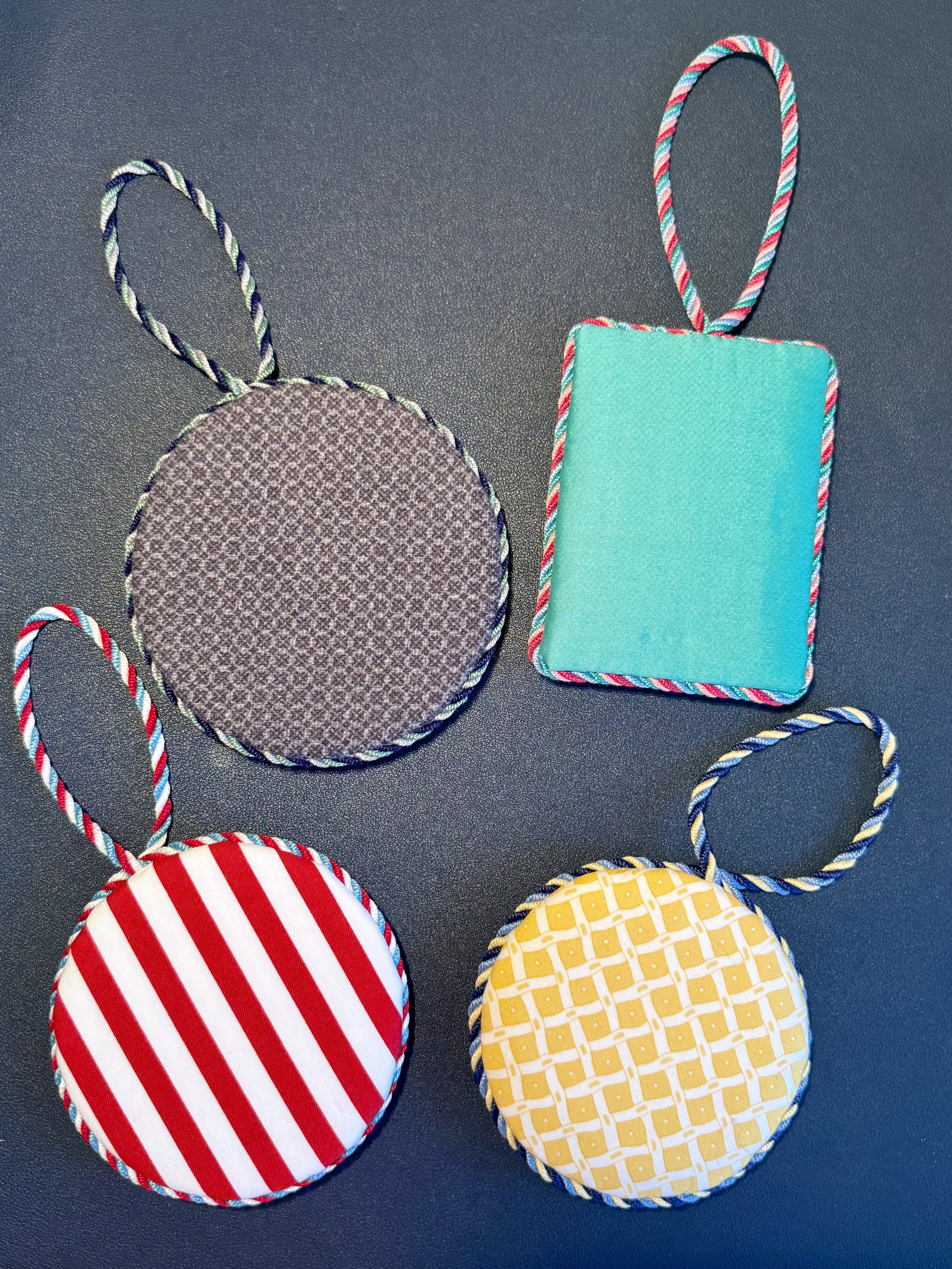 Four fabric-covered objects with string loops, arranged on a dark surface. Two are round with striped pattern, one is square with a plain teal color, and the fourth is partially visible with a yellow and white checkered pattern.