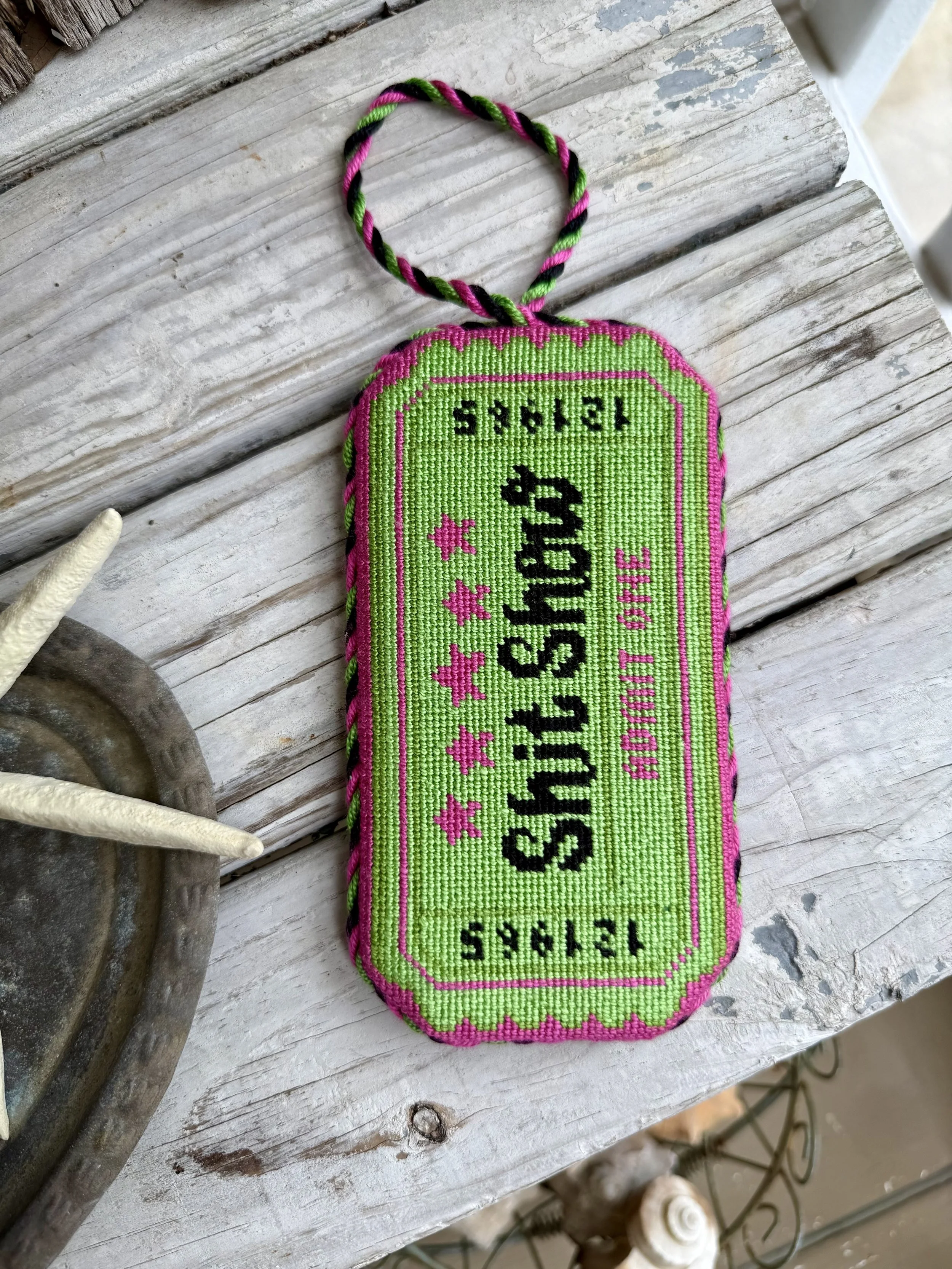 A colorful  finished needlepoint ornament with custom cording, resembling a ticket with pink, green, and black thread. Shit Show Ticket hand stitched,  Finished with The Salty Stitchery