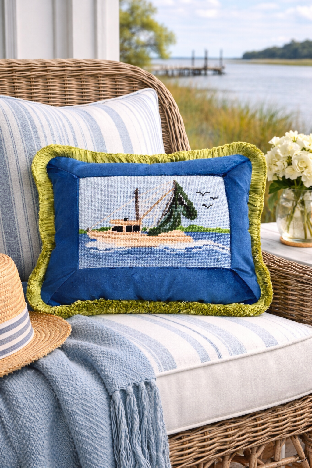 Decorative pillow with a boat, tree, and birds on a body of water, placed on a wicker outdoor sofa with striped cushions, next to a hat, a blue throw blanket, and a glass vase of white flowers, overlooking a river and a wooden dock.