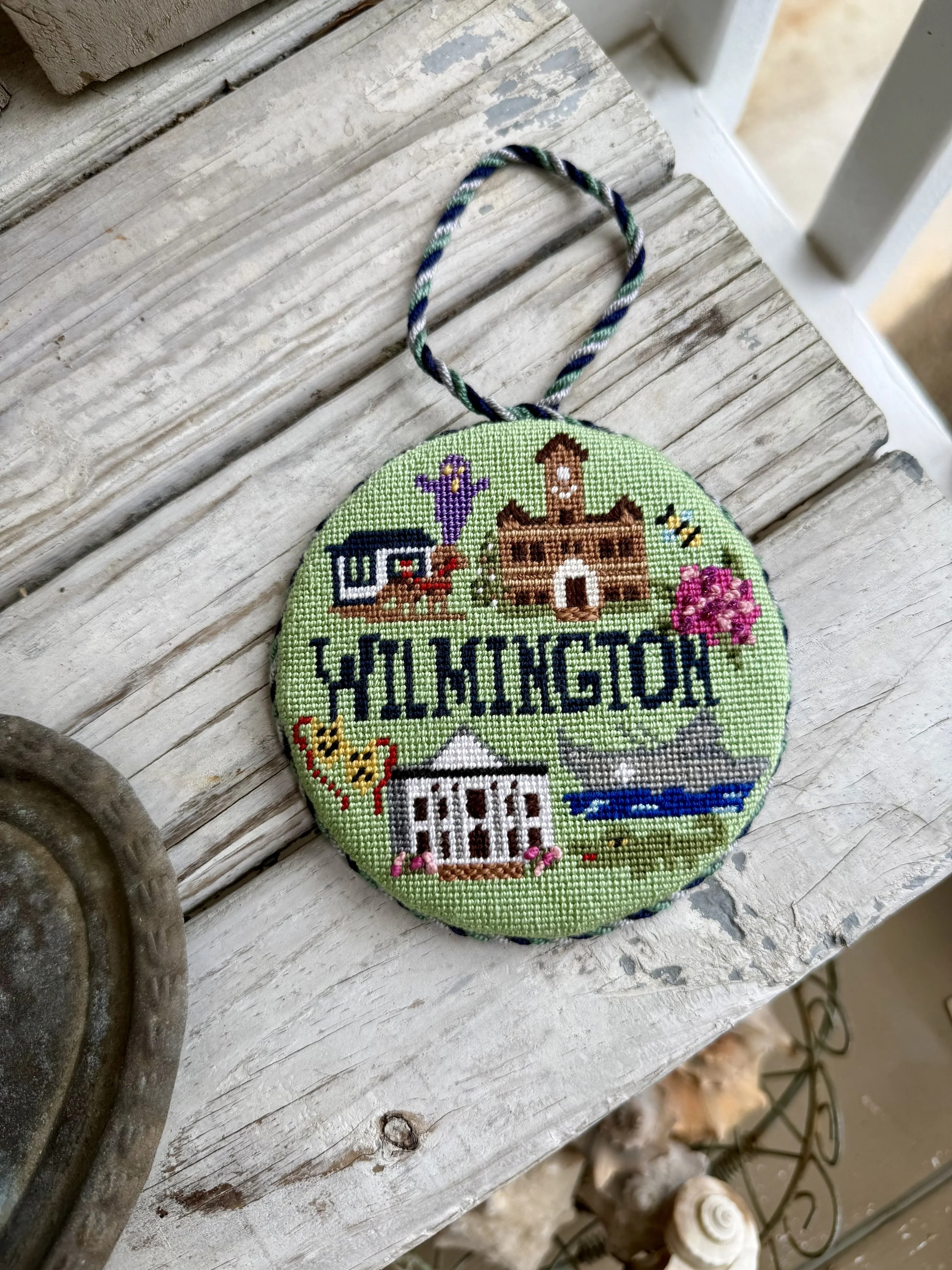 Round, finished needlepoint ornament with a custom cording loop, featuring a house, flowers, and the word "Wilmington" on a green background,  Finished with The Salty Stitchery