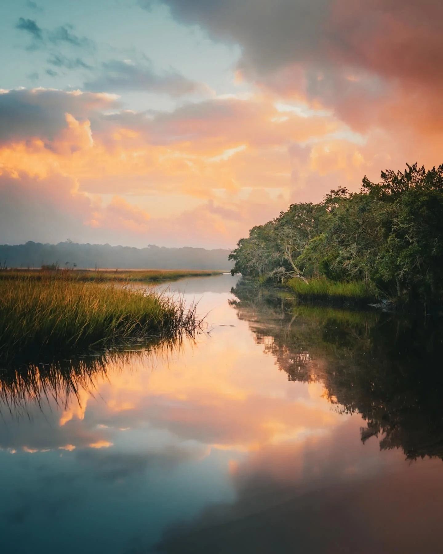 Sunset over a calm river with colorful clouds and dense green trees along the riverbank.