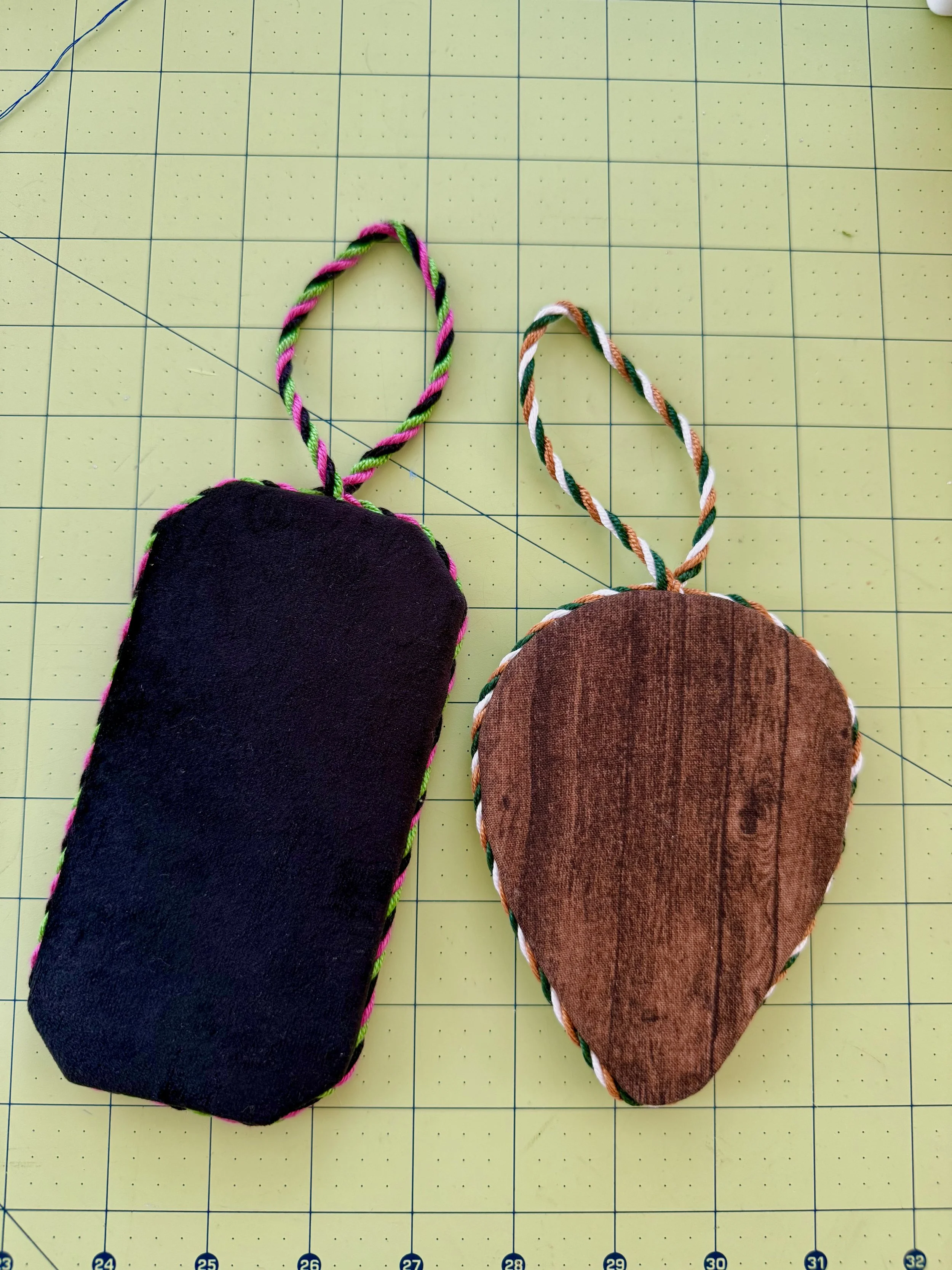 Two needlepoint ornaments connected by a multicolored string, one black and one brown, placed on a yellow grid cutting mat.