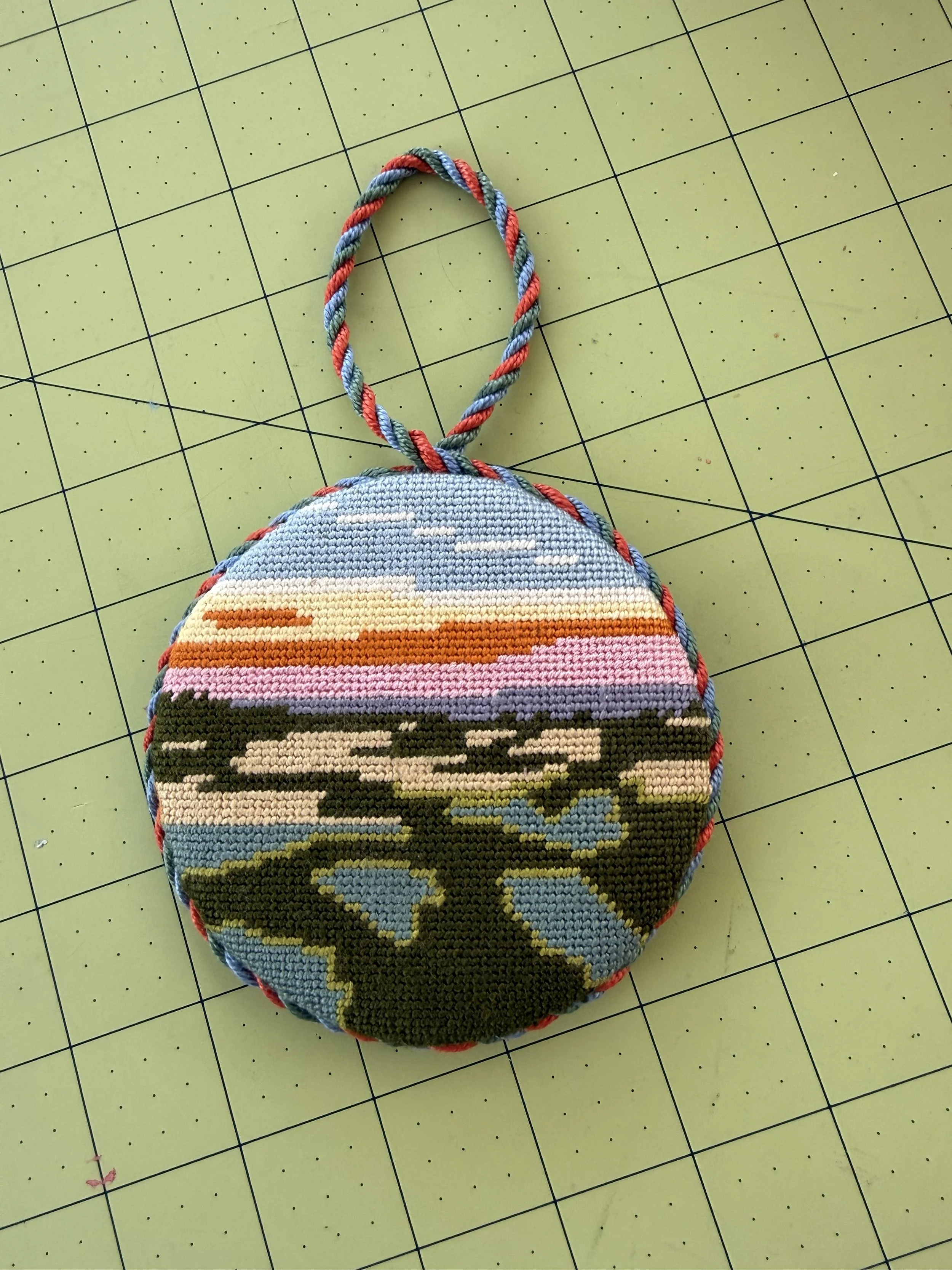 A round, needlepoint item with a multi-colored design and a twisted loop handle, placed on a green cutting mat with grid lines.