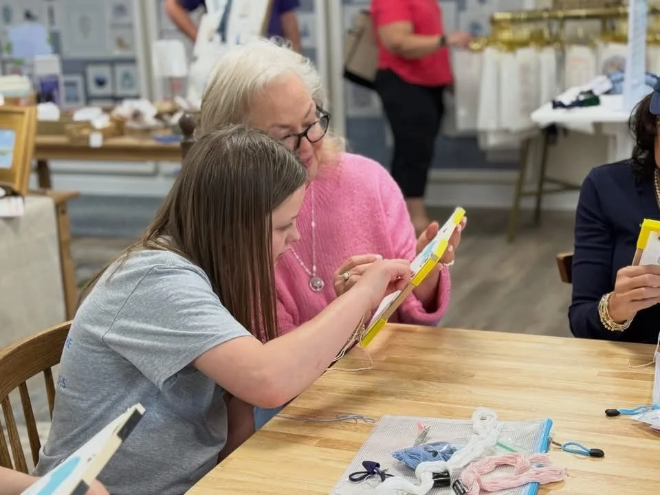 A young girl and a woman sit at a wooden table in a craft store, learning needlepoint together. Other people are seen in the background, browsing and shopping.