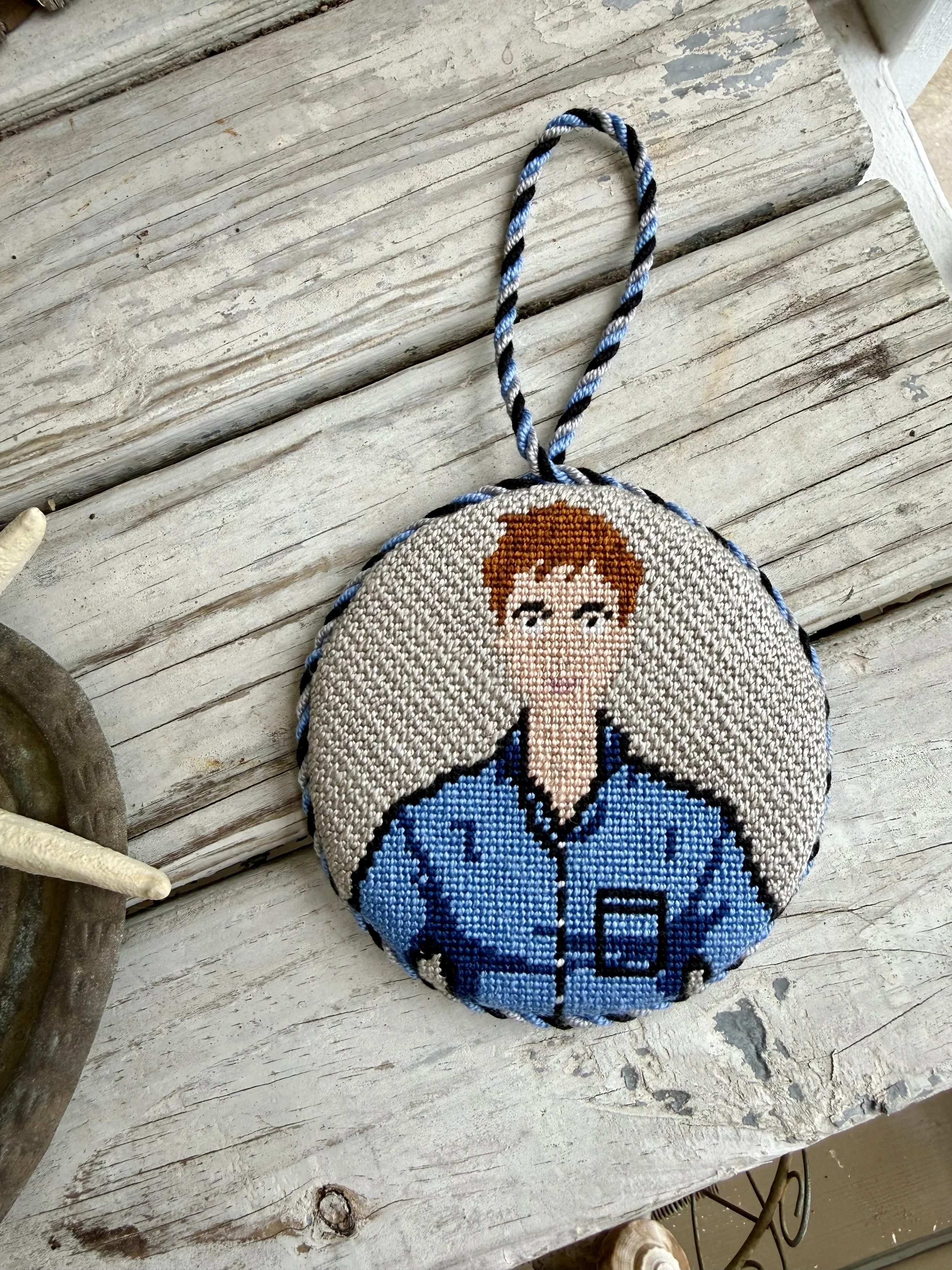 A circular finished needlepoint ornament with a custom cording loop featuring a cartoon face of a person with brown hair, blue eyes, and a blue shirt, on a wooden surface.Finished with The Salty Stitchery
