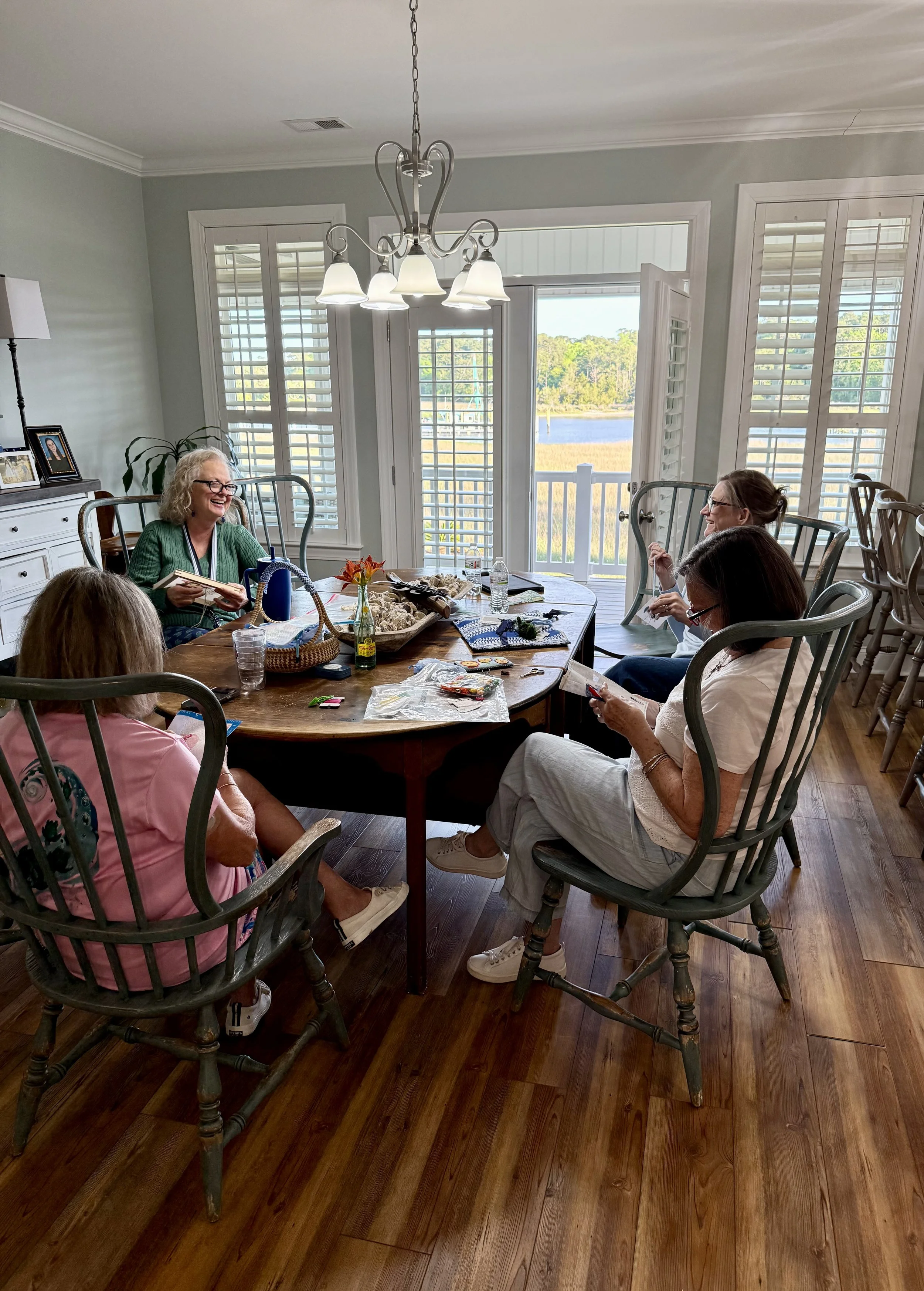 A group of women sitting at a dining table near a large window with white shutters, enjoying conversation; the table has various items, including needlepoint, and a flower in a small vase.