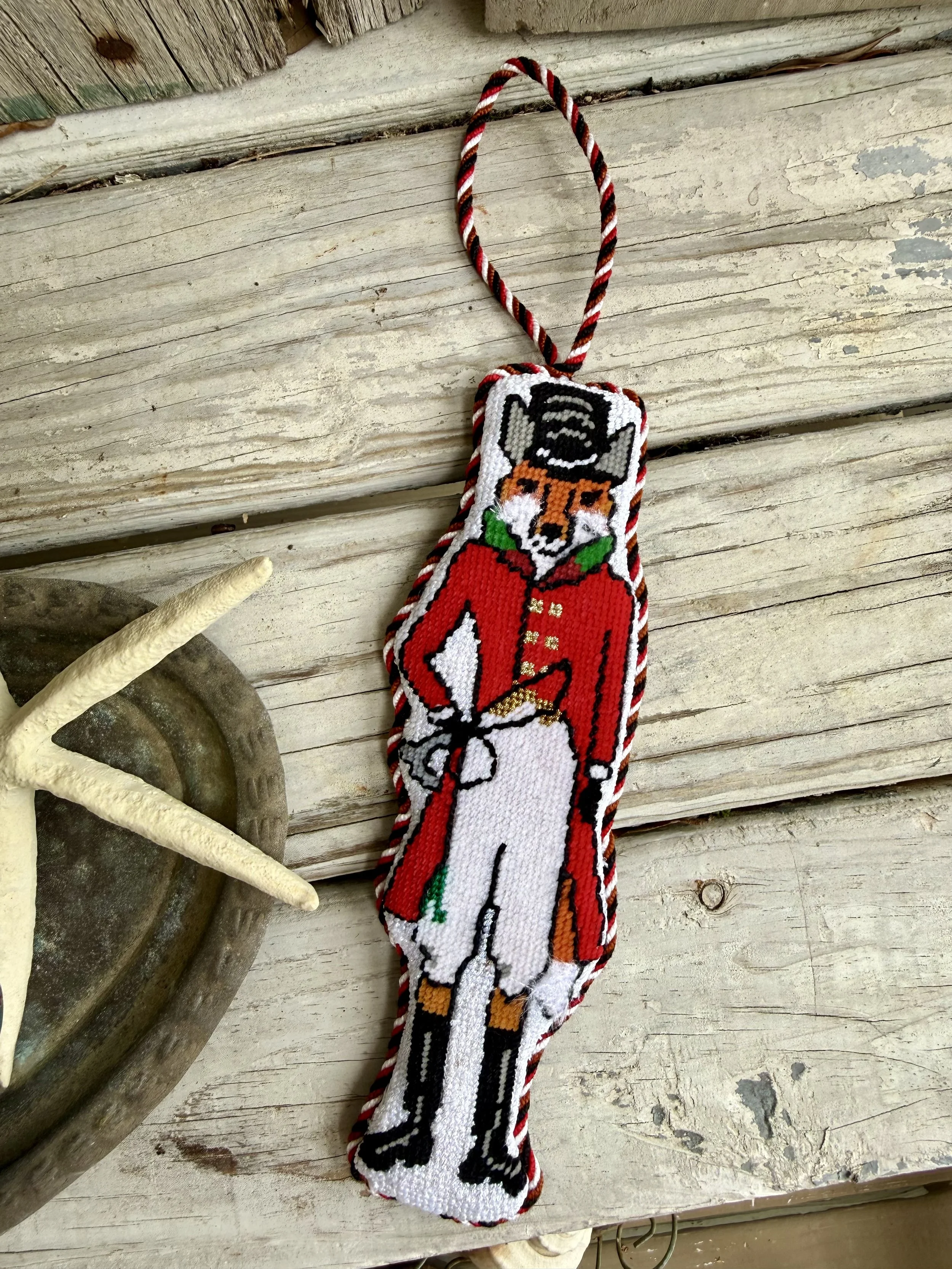 A handmade finished needlepoint ornament with custom cording, featuring a fox wearing a top hat, with red, white, black, and orange colors,  Finished with The Salty Stitchery