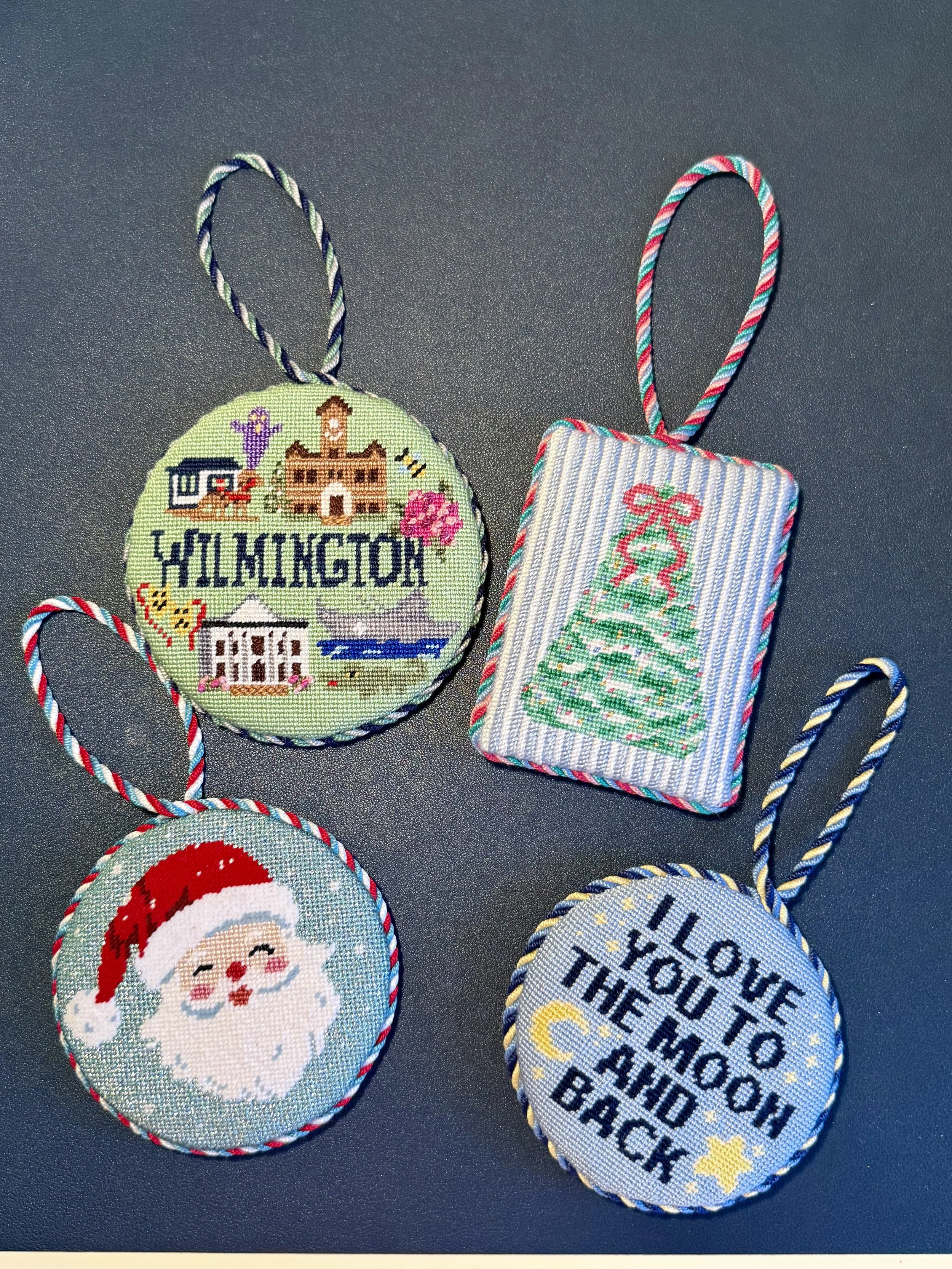 Needlepoint Christmas ornaments, including a round ornament with the town of Wilmington, a rectangular ornament with a Christmas tree, and an ornament with Santa Claus's face, all with colorful striped borders.