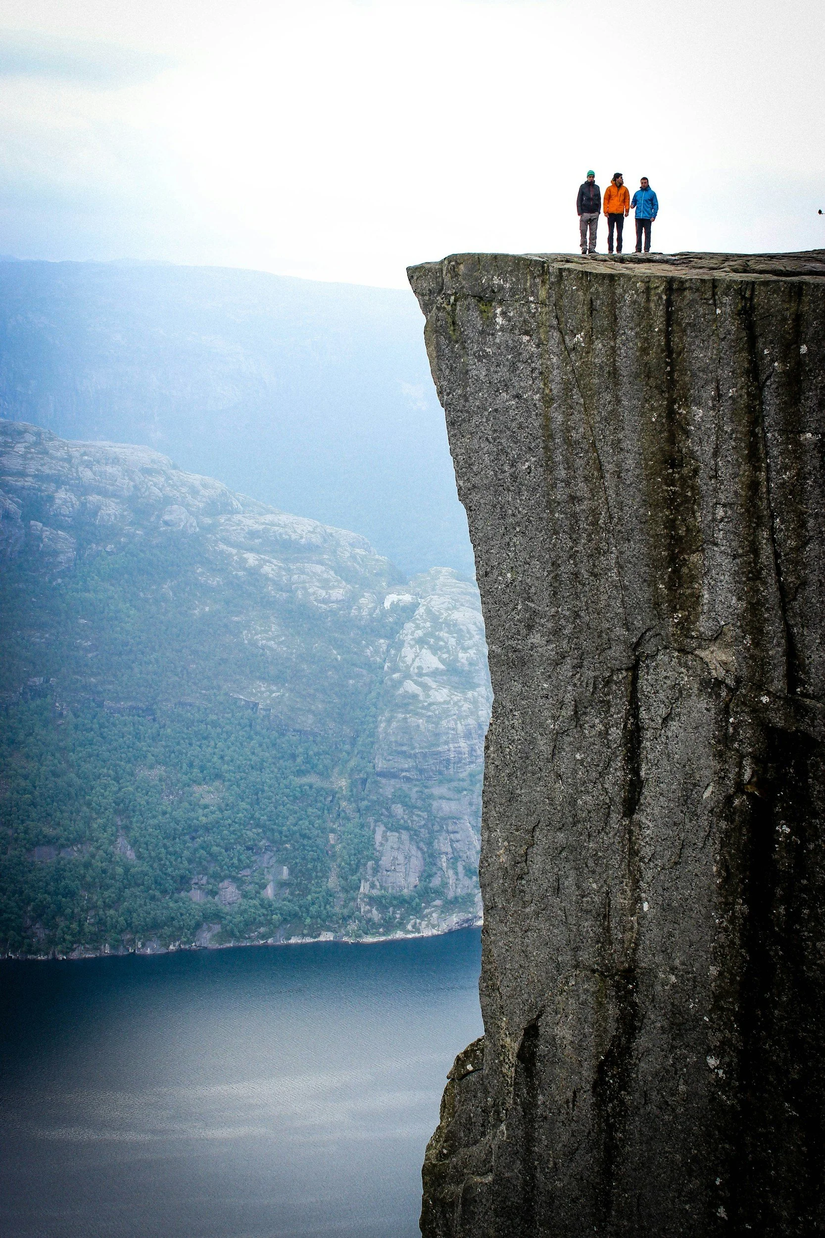 Three people stand at the edge of a steep cliff overlooking a body of water and distant mountains in a foggy landscape.