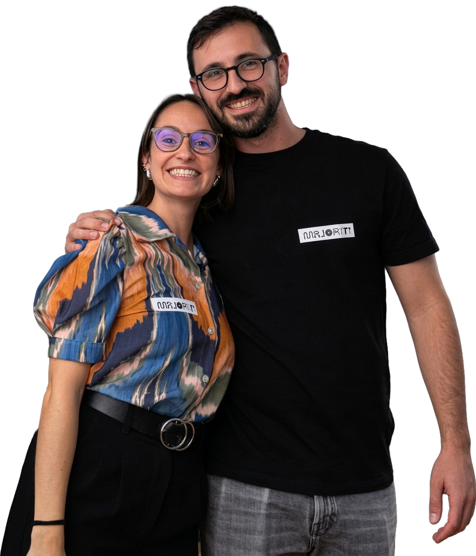 A smiling woman and man with glasses, embracing each other. The woman has short brown hair and is wearing a colorful, patterned blouse with a black belt. The man has dark hair, a beard, and is wearing a black T-shirt and gray pants. Both are wearing name tags.