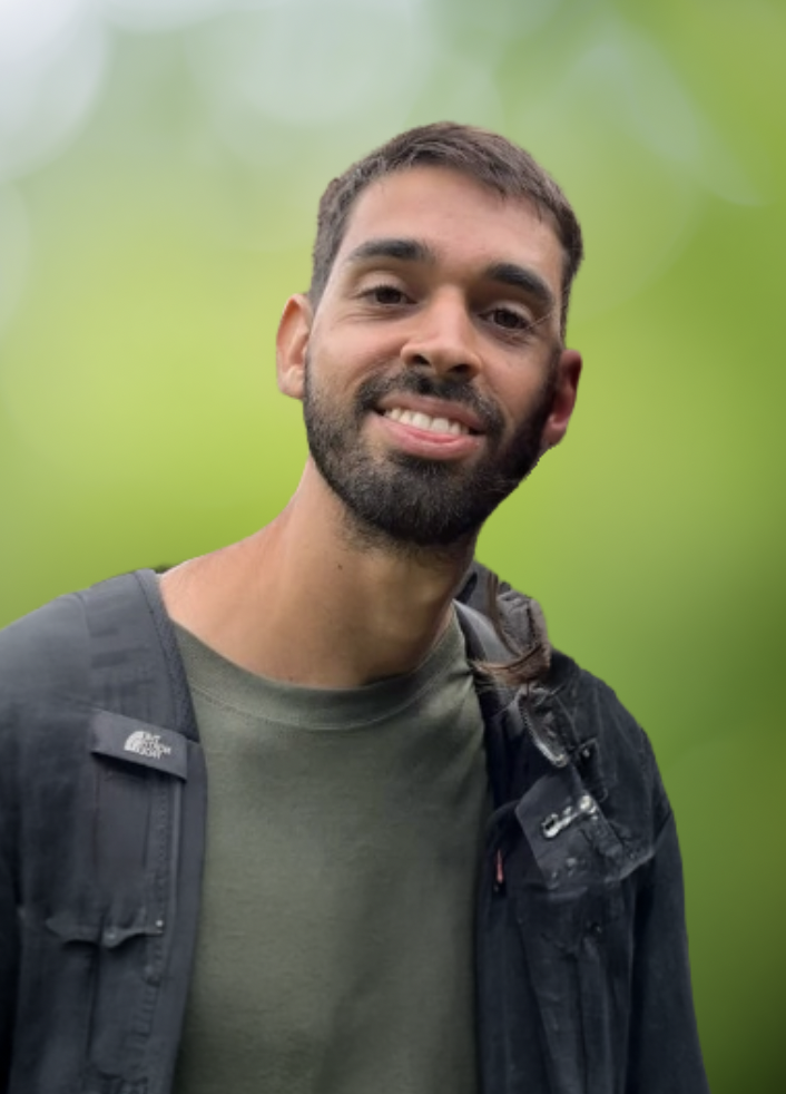 A man outdoors with a blurred green background, wearing a dark jacket and a green shirt, smiling and carrying a backpack.