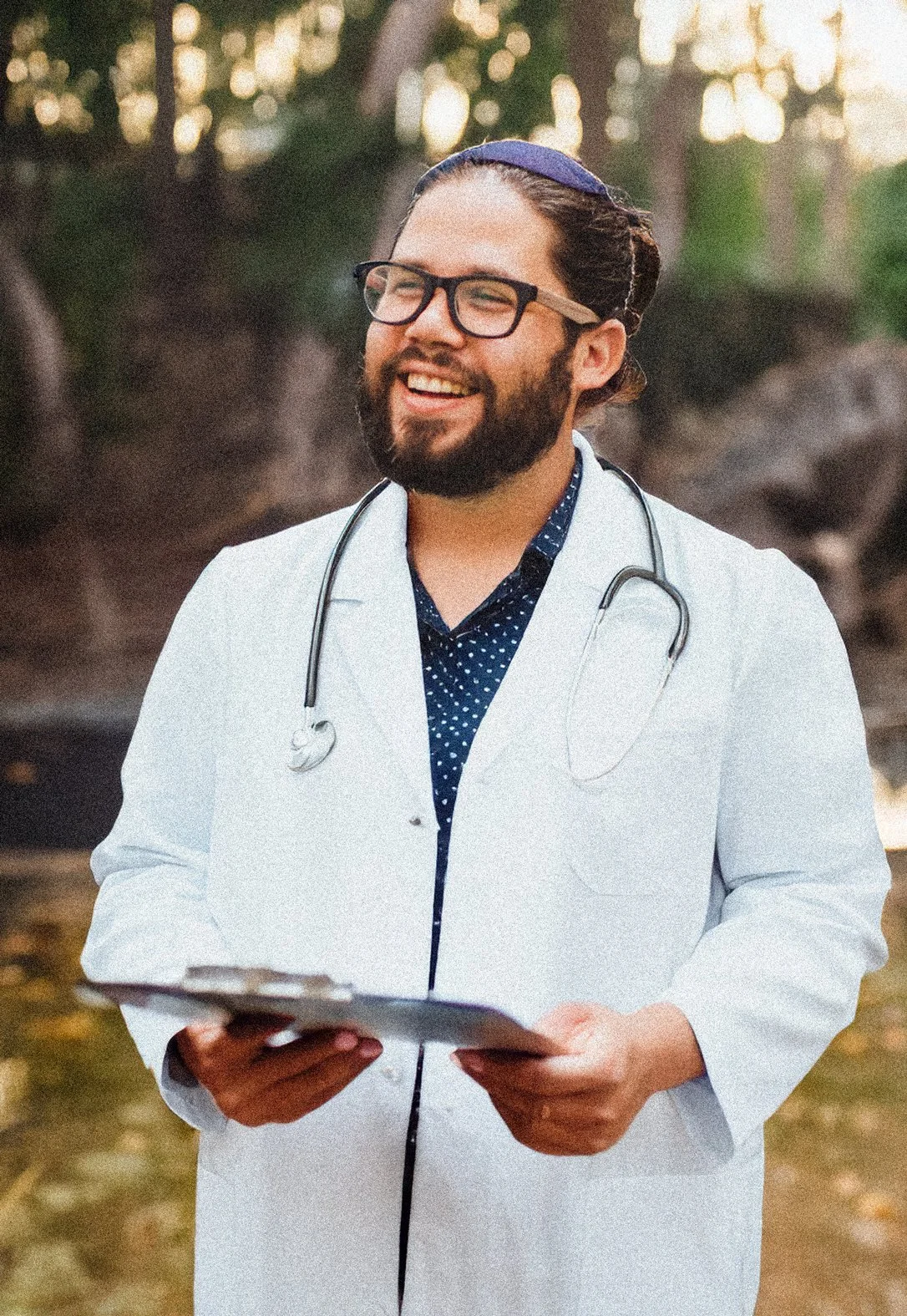 A male doctor with glasses and a beard, wearing a white coat and stethoscope, holding a clipboard outdoors during daytime, smiling.