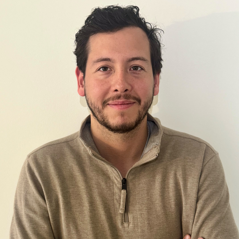 A young man with dark, curly hair, a slight beard, and a mustache, smiling and looking at the camera, wearing a beige quarter-zip pullover sweater in front of a plain white wall.