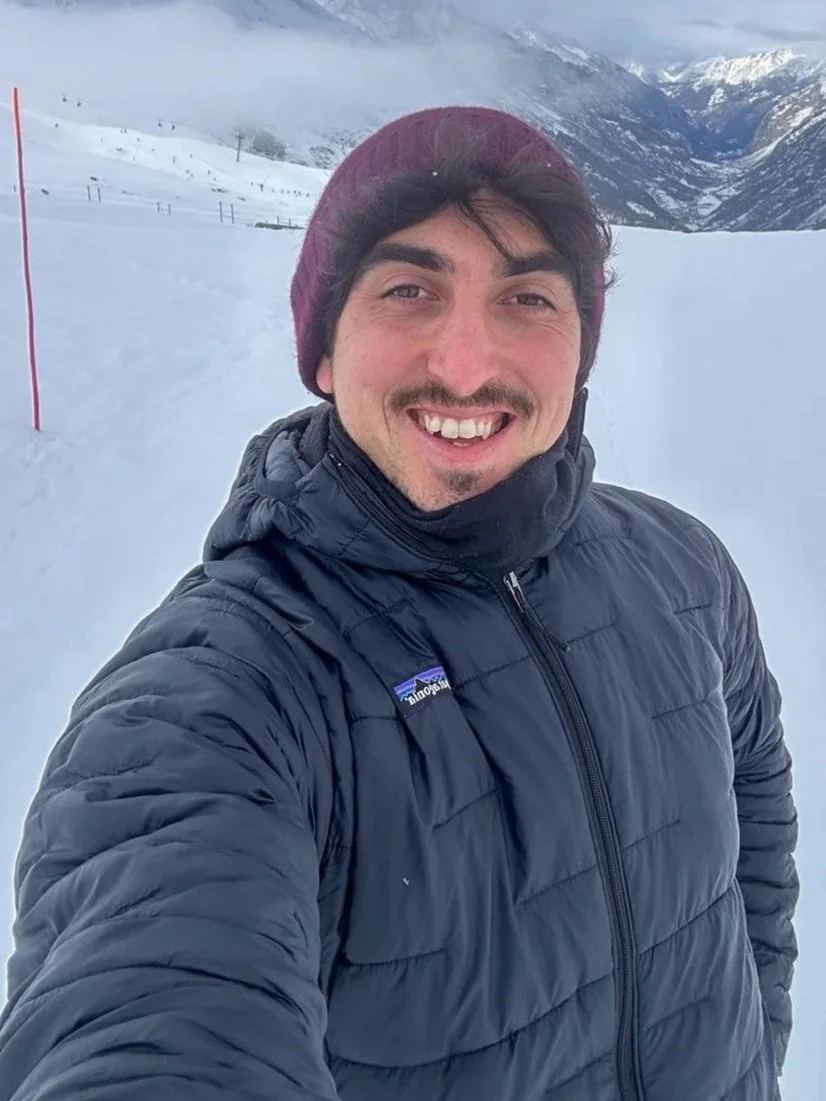 A man smiling for a selfie in a snowy mountainous landscape, wearing a dark puffy jacket, a maroon beanie, and a neck gaiter.