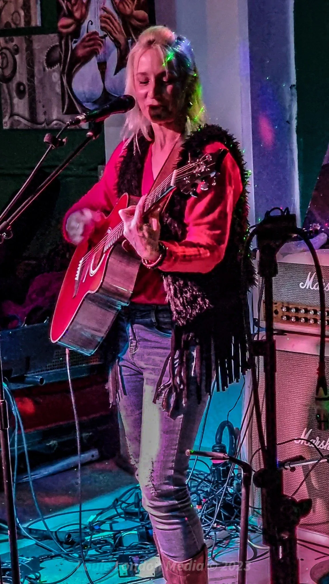 Femme jouant de la guitare électrique en concert dans une ambiance colorée avec des amplificateurs et des câbles.