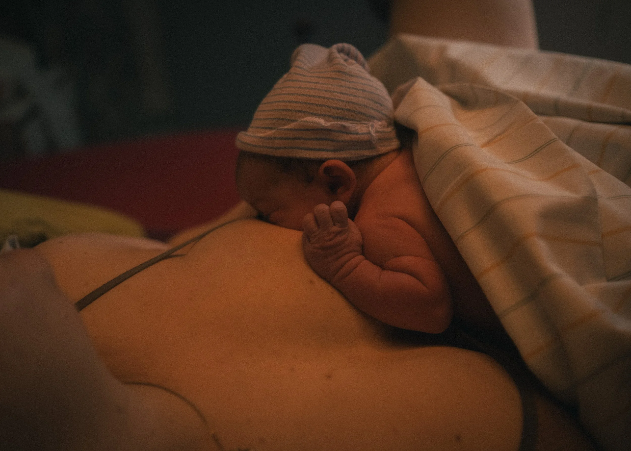 Ein Neugeborenes, das auf der Brust einer Person liegt, trägt eine gestreifte Mütze und ist mit einer Decke bedeckt.