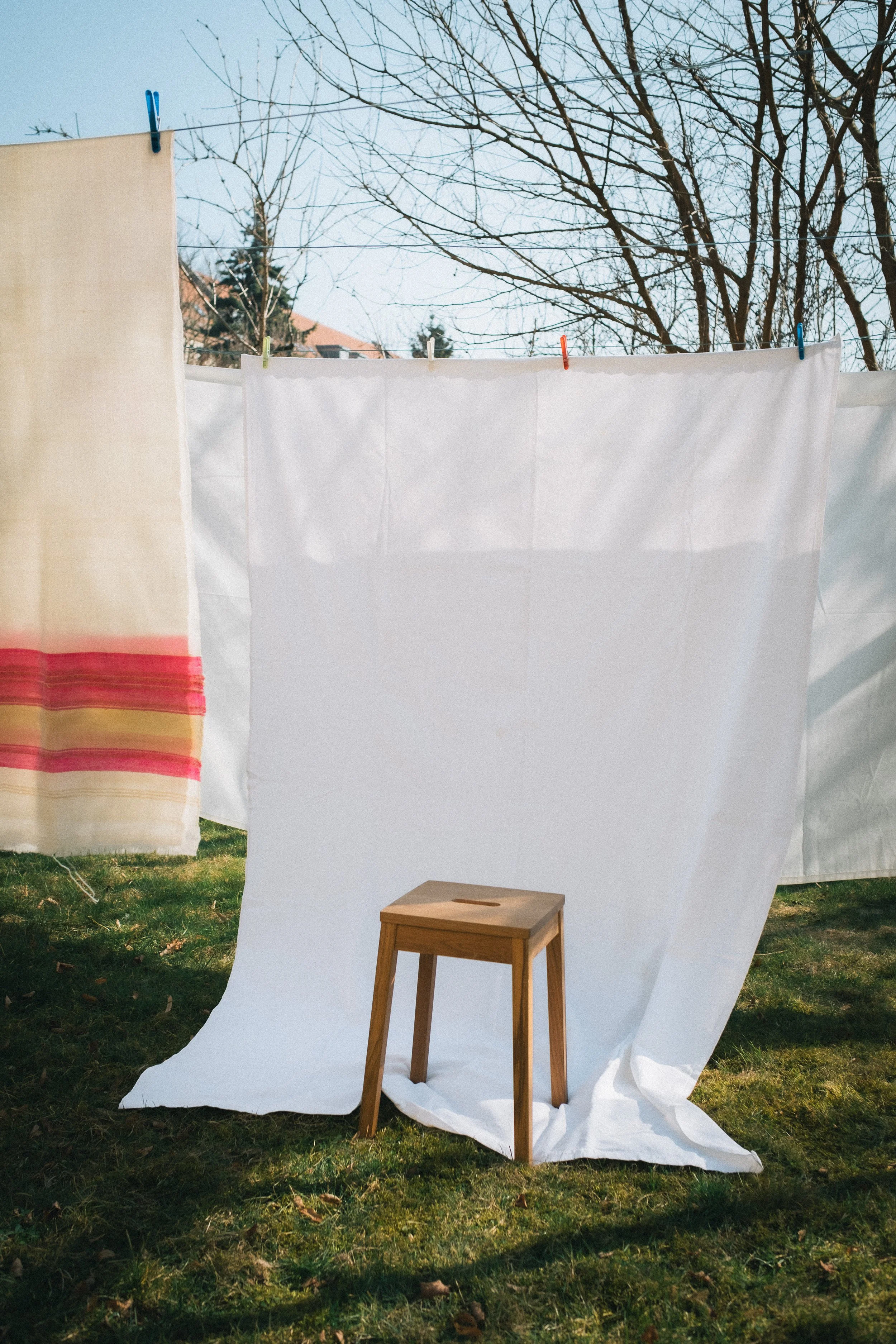 Weiße Wäsche hing auf einer Wäscheleine im Garten, mit einem kleinen Holzhocker auf dem gras. Der Himmel ist blau, Bäume ohne Blätter sind im Hintergrund.