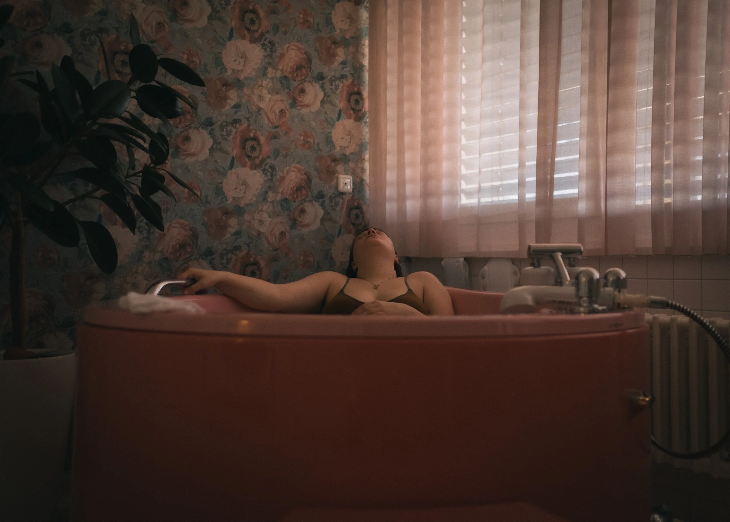 Frau liegt entspannt in einer Badewanne, eingerahmt von einer floral gestalteten Tapete und einem weißen Vorhang, in einem badezimmerartigen Raum.