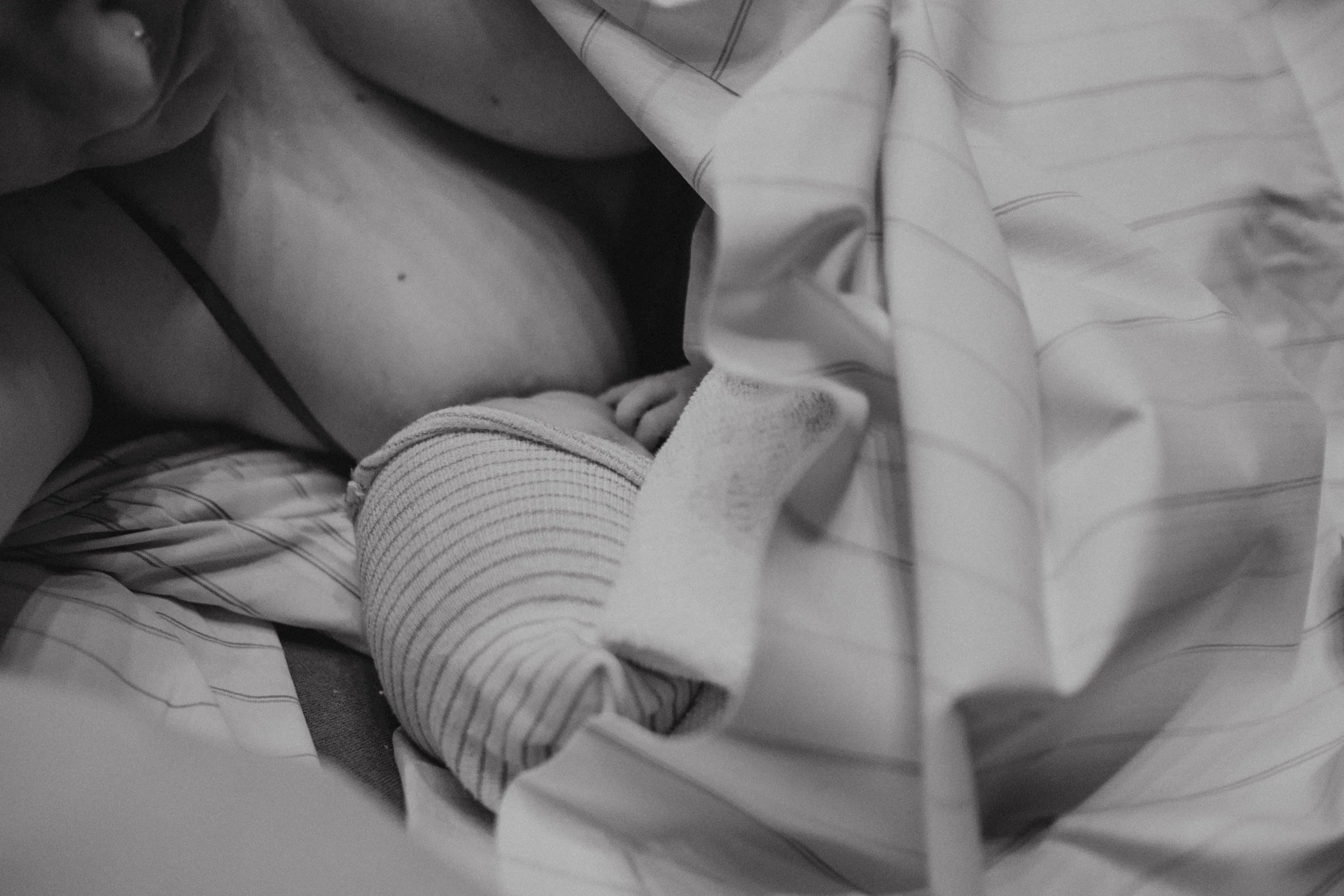 Neugeborenes Baby, eingewickelt in gestreiftes Tuch, liegt Bauch an Bauch mit der Mutter und wird gestillt.