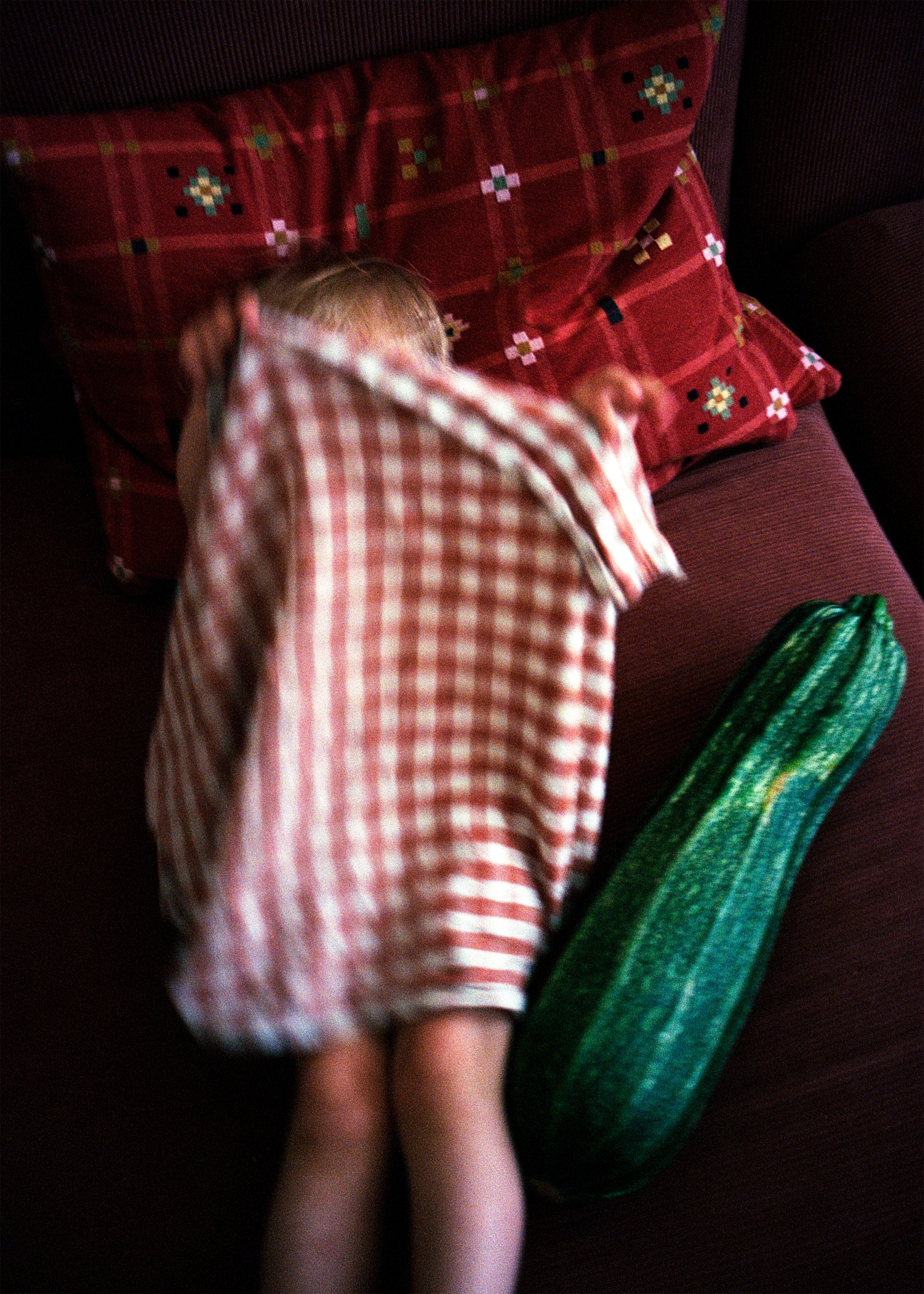 Ein Kind liegt schlafend auf einem roten Sofa, bedeckt mit einem rot-weiß karierten Tuch, mit roten Kissen im Hintergrund und einem langen grünen Kissen auf der Seite.