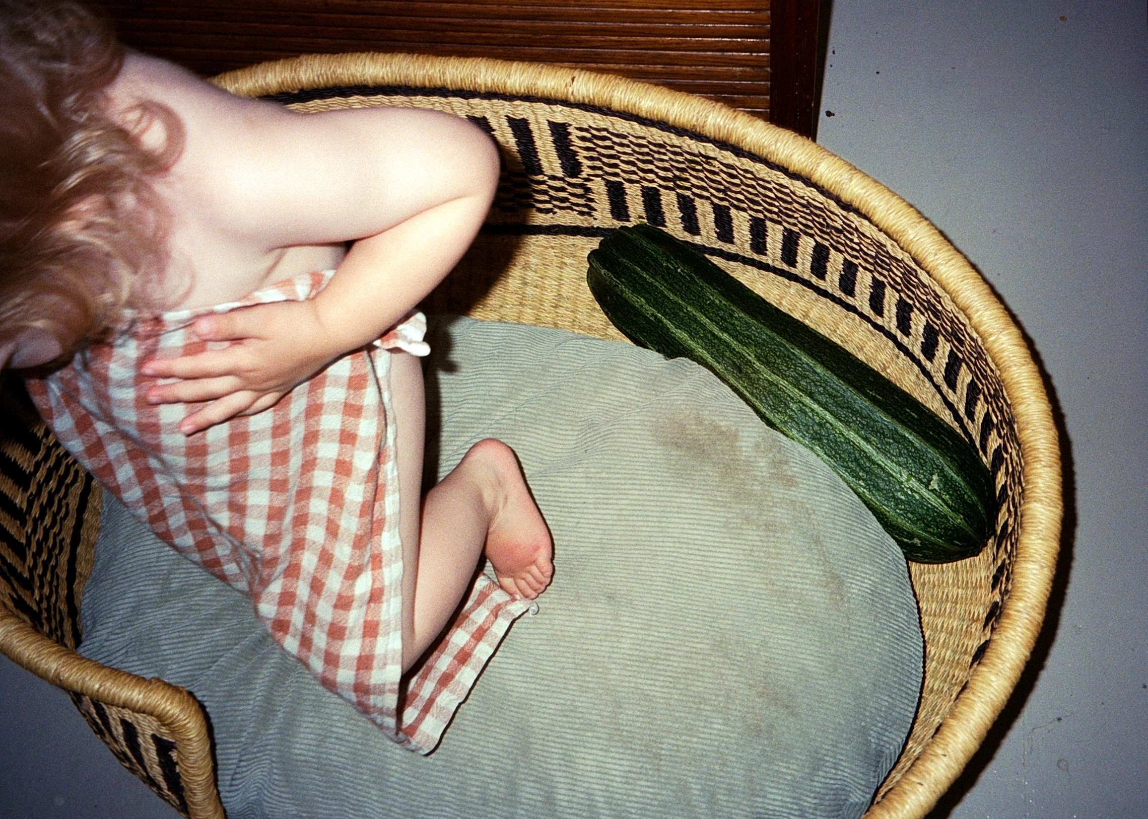 Ein kleiner Junge sitzt auf einem Korbstuhl, halb bedeckt mit einem rot-weißen karierten Handtuch, und liegt neben einem großen Zucchini.