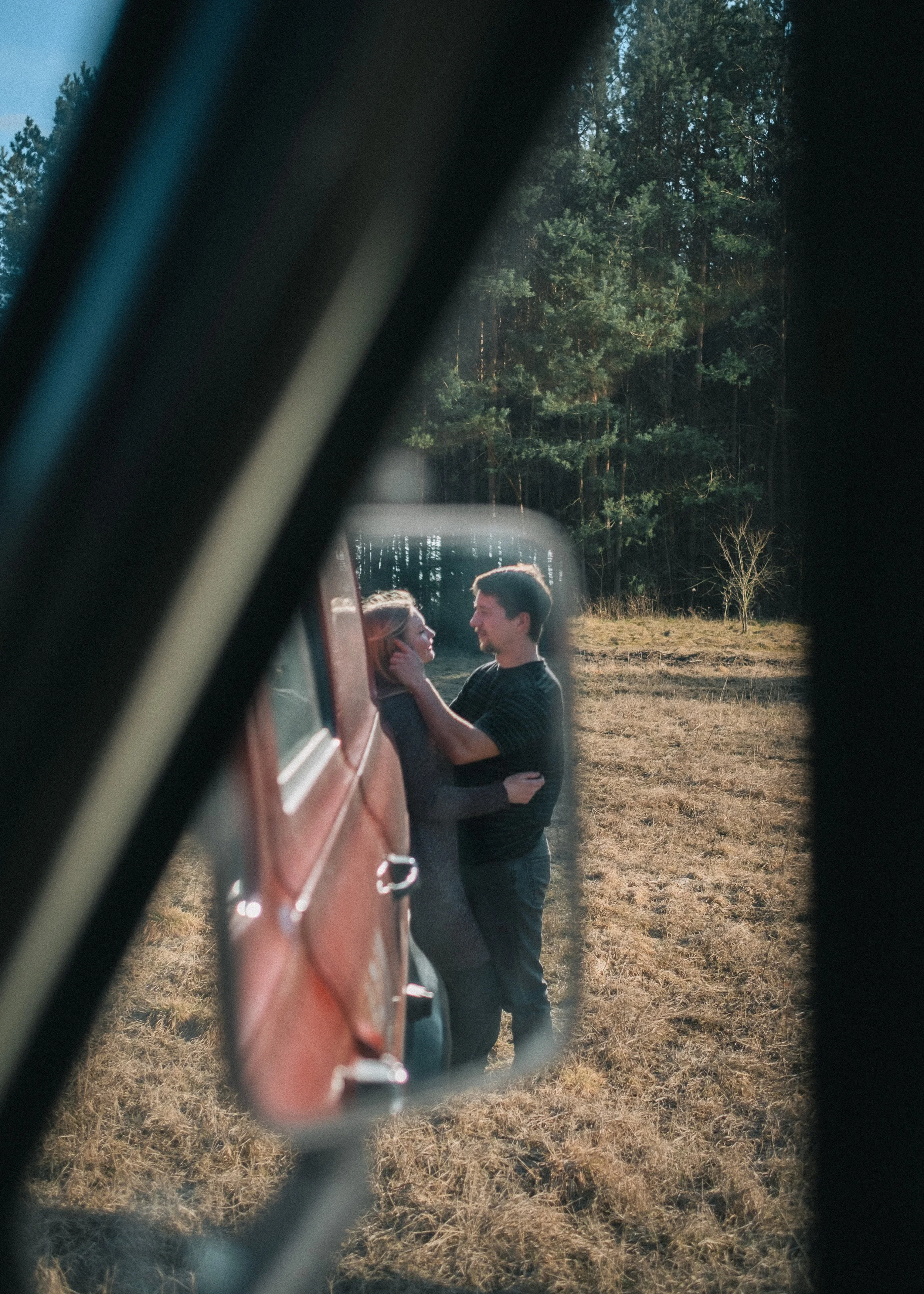 Zwei Menschen, ein Mann und eine Frau, sehen sich in Nahaufnahme durch den Außenspiegel eines Autos an, während sie draußen auf einem Feld mit Bäumen im Hintergrund stehen.
