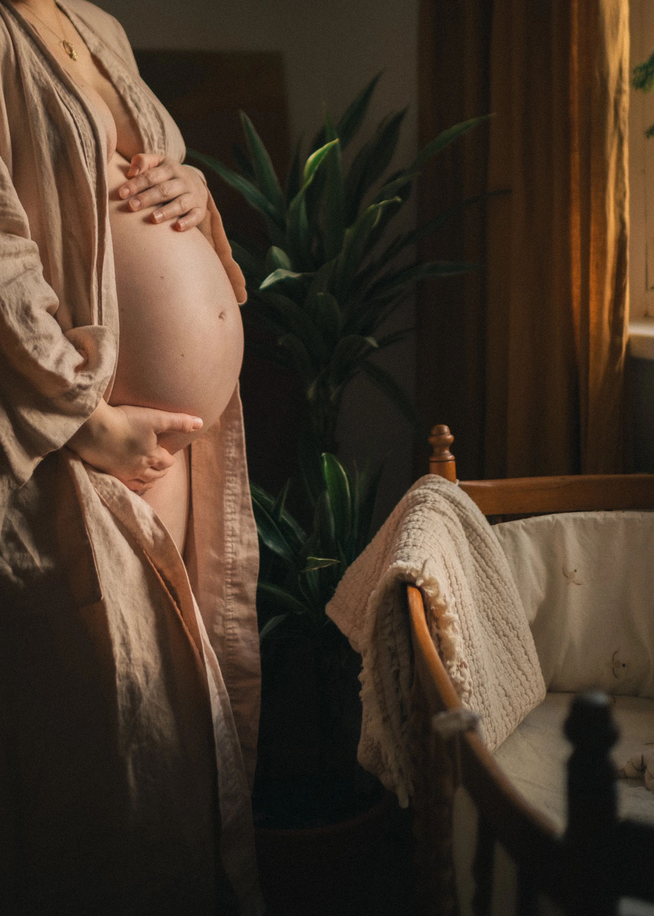 Eine schwangere Frau steht in einem Raum, hält ihren Babybauch mit einer Hand und trägt einen beiläufigen Kimono. Im Hintergrund sind Pflanzen und ein Sofa mit einer gestrickten Decke. Das Licht ist warm und weich.