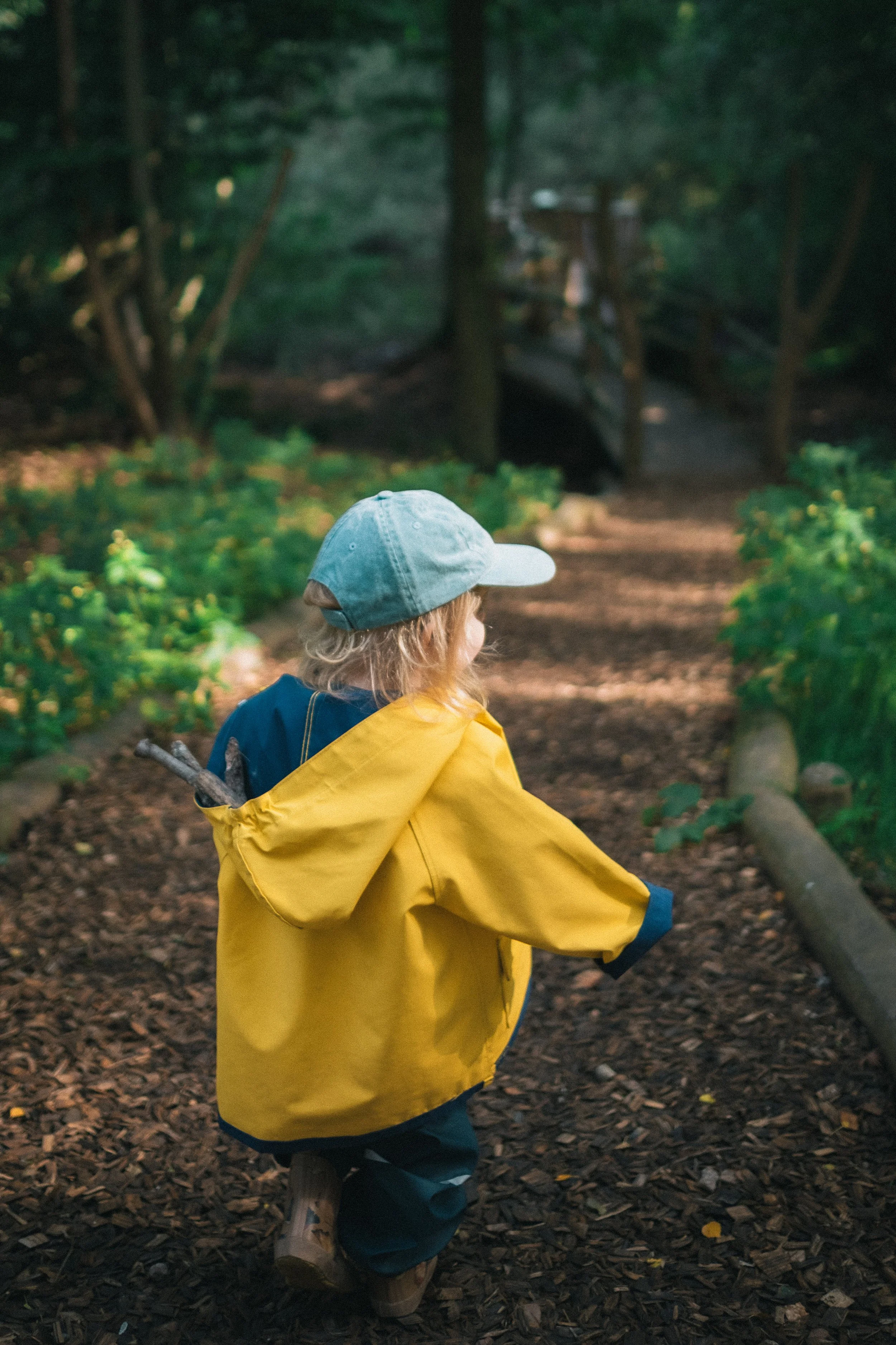 Ein Junge in gelber Regenjacke und Mütze läuft auf einem Waldweg. Im Hintergrund ist eine kleine Brücke zu sehen.