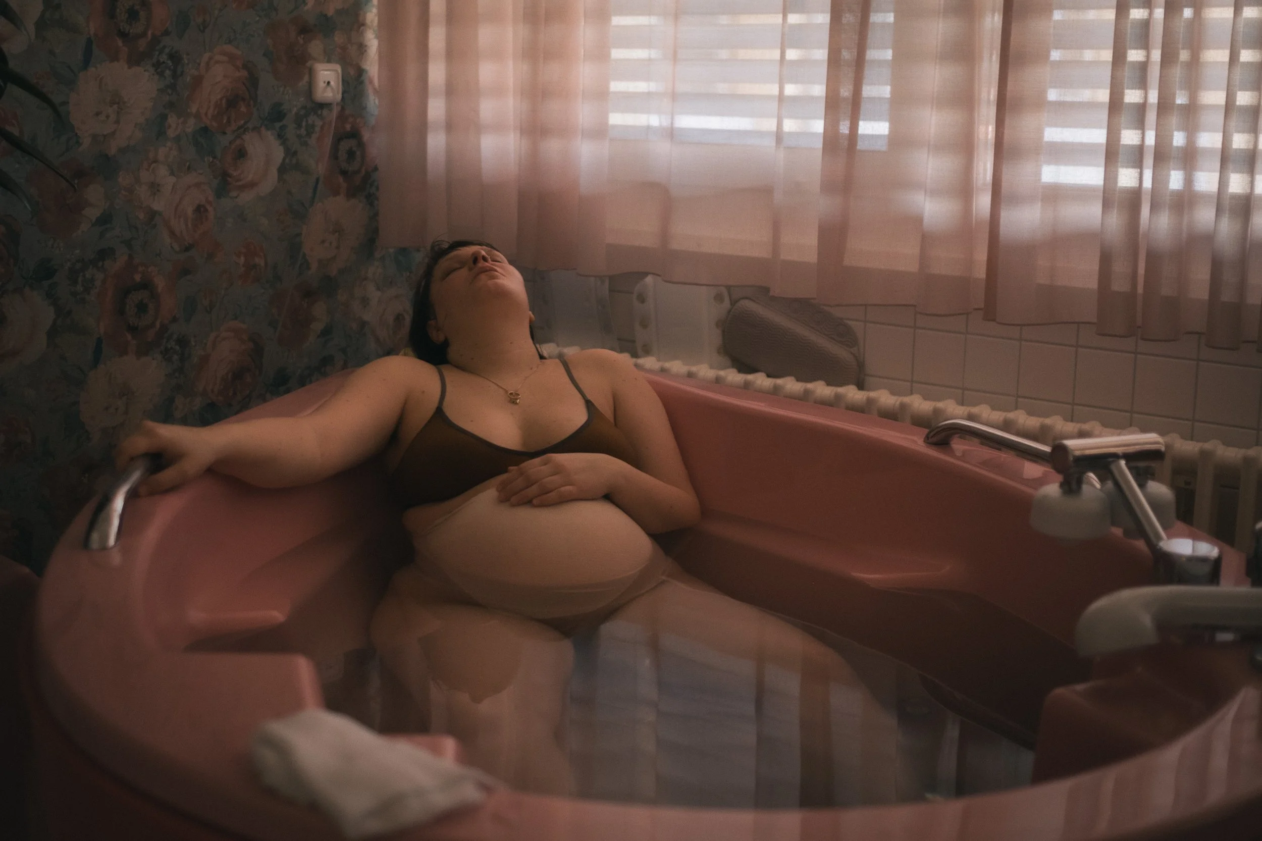 Frau liegt im  verdunkelten Kreissal in einer rosanen Geburtswanne den Kopf nach hinten gelegt und eine Hand auf dem Bauch.