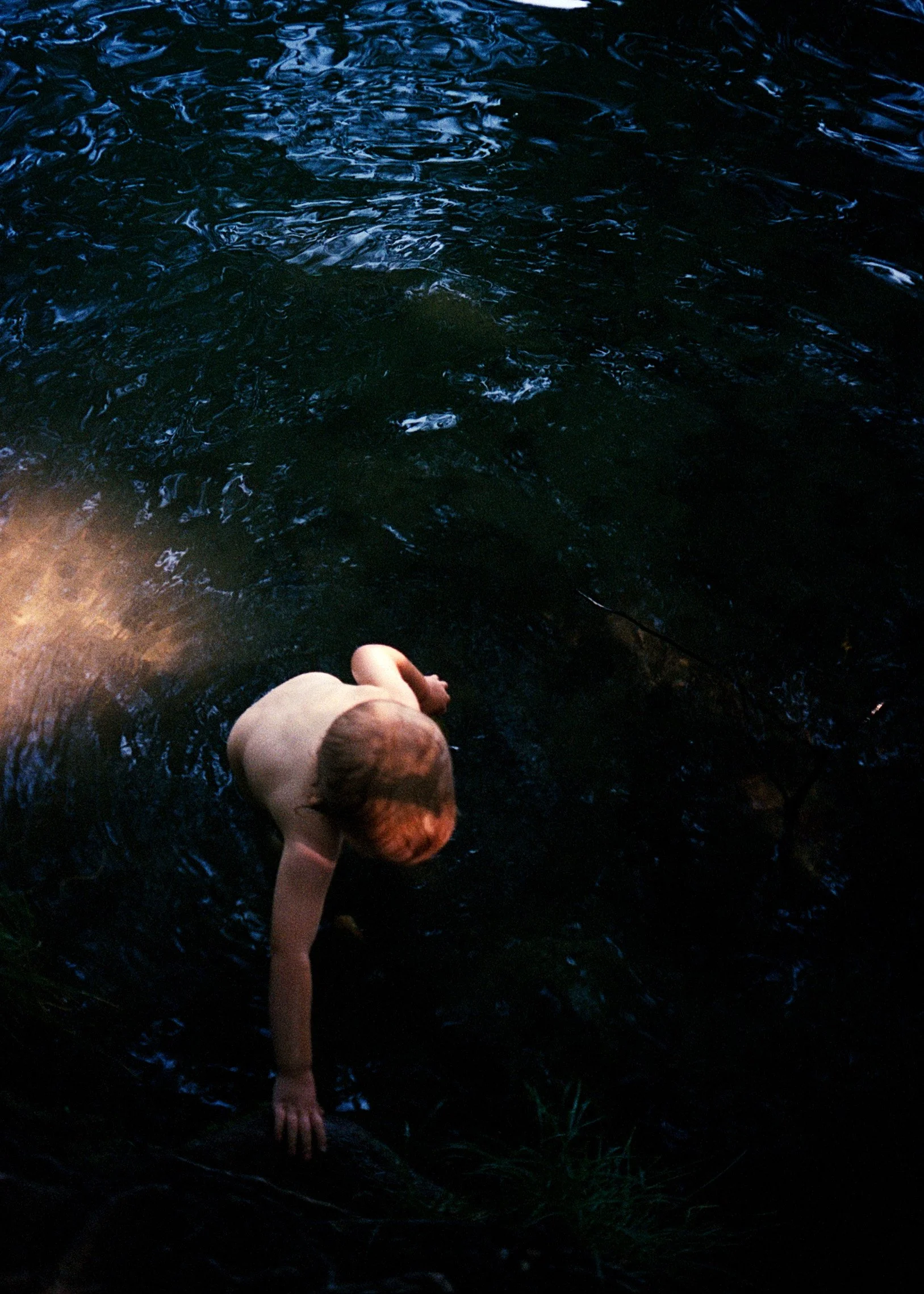 Ein nackter Junge sitzt an einem Flussufer im Dämmerlicht und schaut ins Wasser.