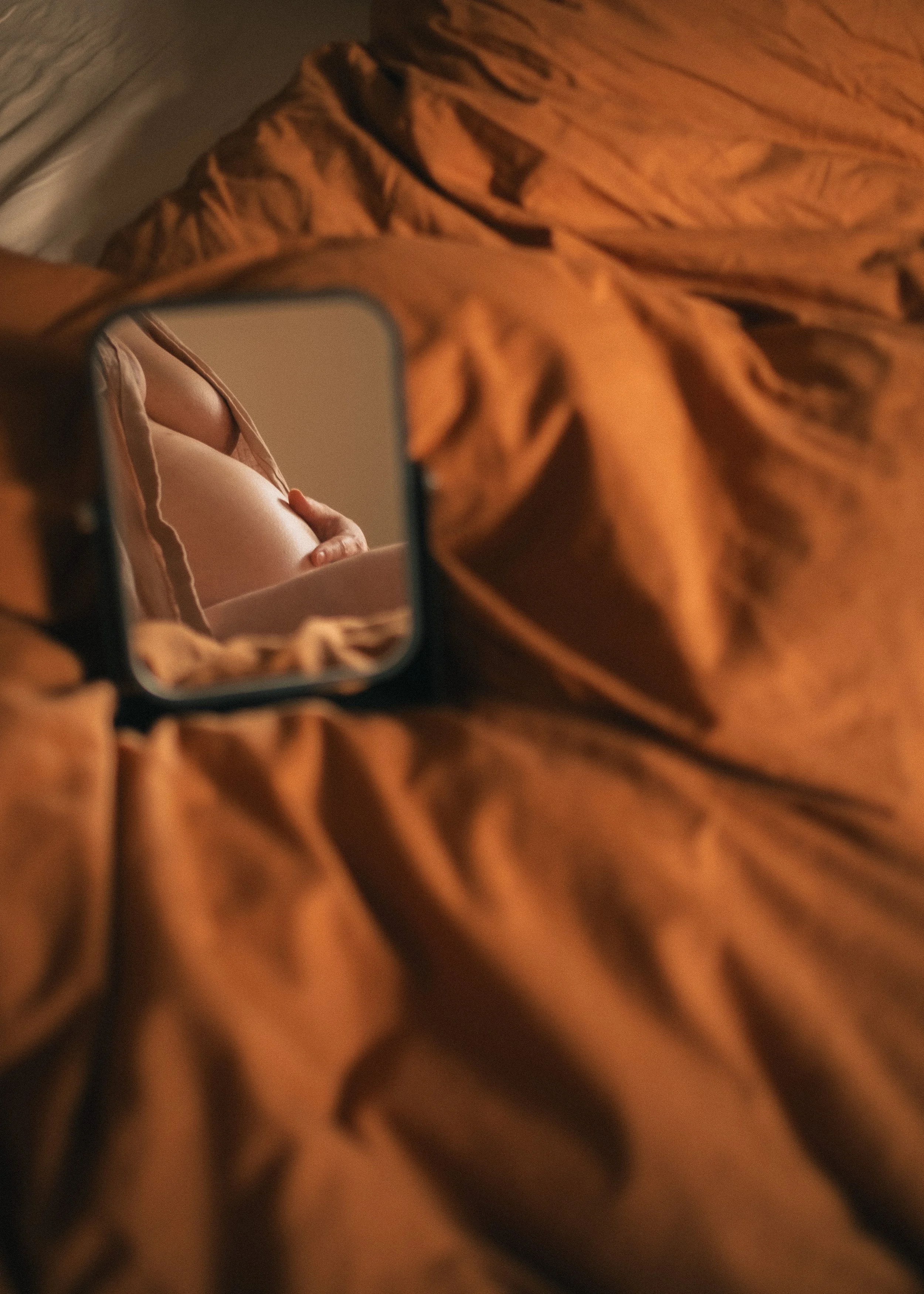 Nahaufnahme eines Mannes, der in einen kleinen Spiegel schaut, der auf einem braunen, altmodischen Bettdecke liegt. Im Spiegel ist nur ein Teil des Oberkörpers eines Mannes sichtbar, der an seinem Bauch berührt.