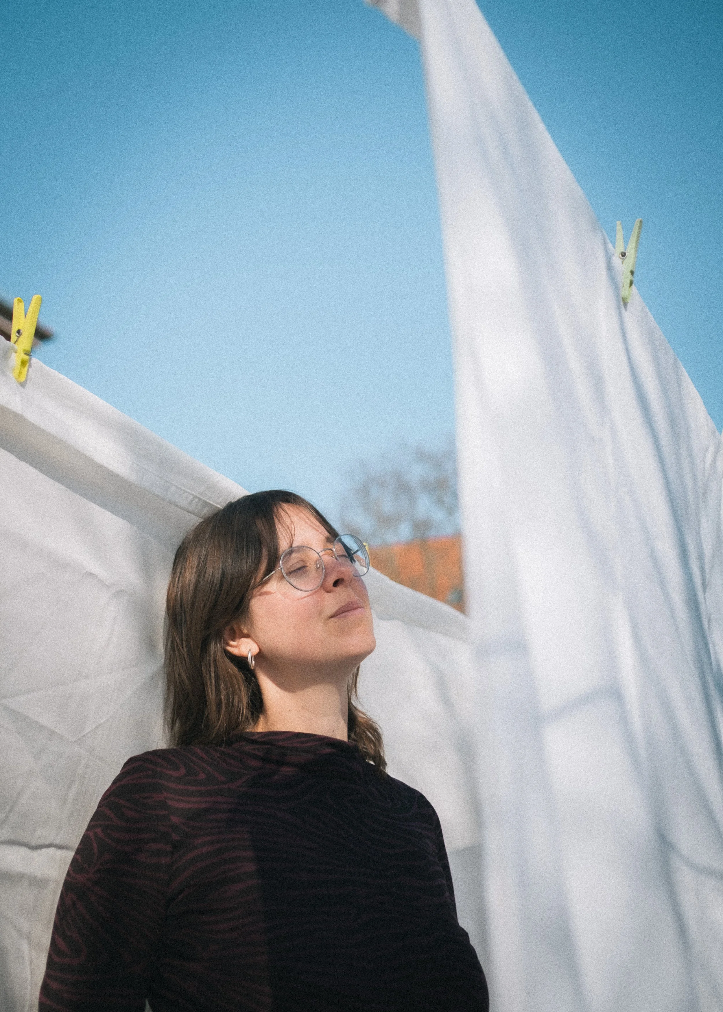 Eine Frau mit Brille und dunklem Oberteil steht vor einem Wäscheleine mit weißen Wäschestücken, die an einem sonnigen Tag gehängt sind.