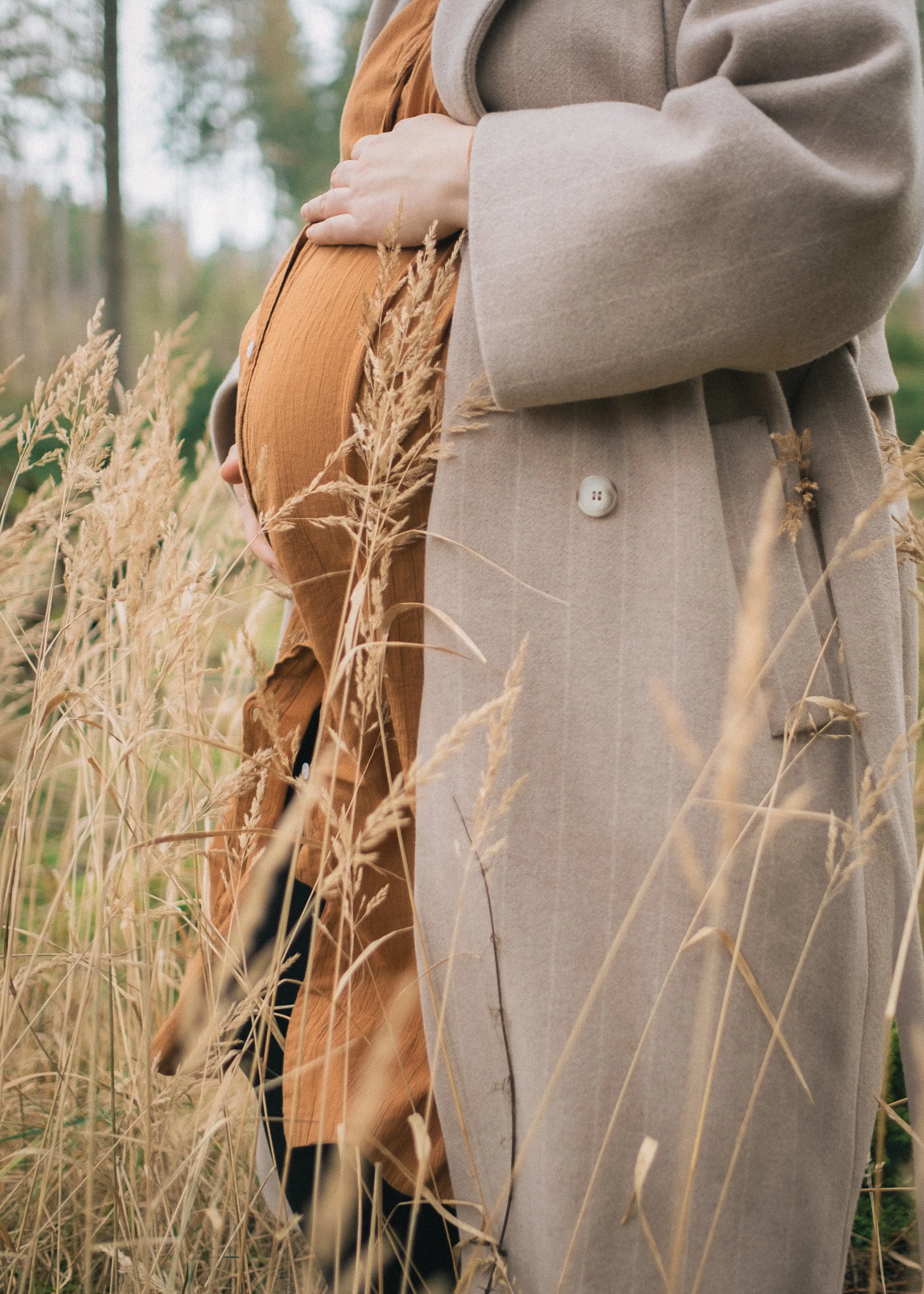 Nahaufnahme eines schwangeren Bauches, gehalten von einer Person in einem beige Mantel, im Freien mit hohem, trockenen Gras im Vordergrund.