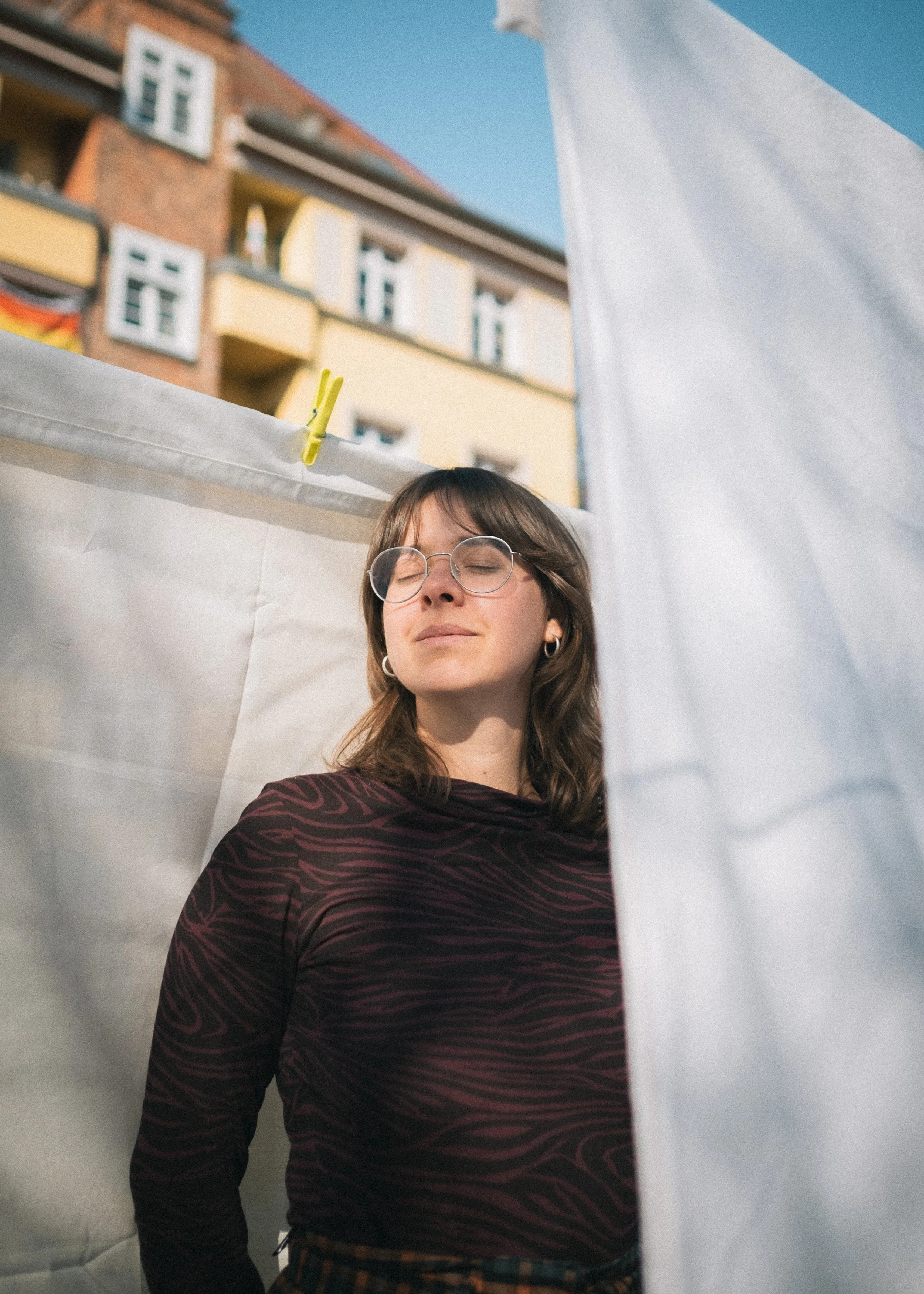 Eine junge Frau mit Brille und Ohrringen steht zwischen weißen Wäscheleinen im Freien in der Sonne, mit einem mehrstöckigen Gebäudekomplex im Hintergrund.