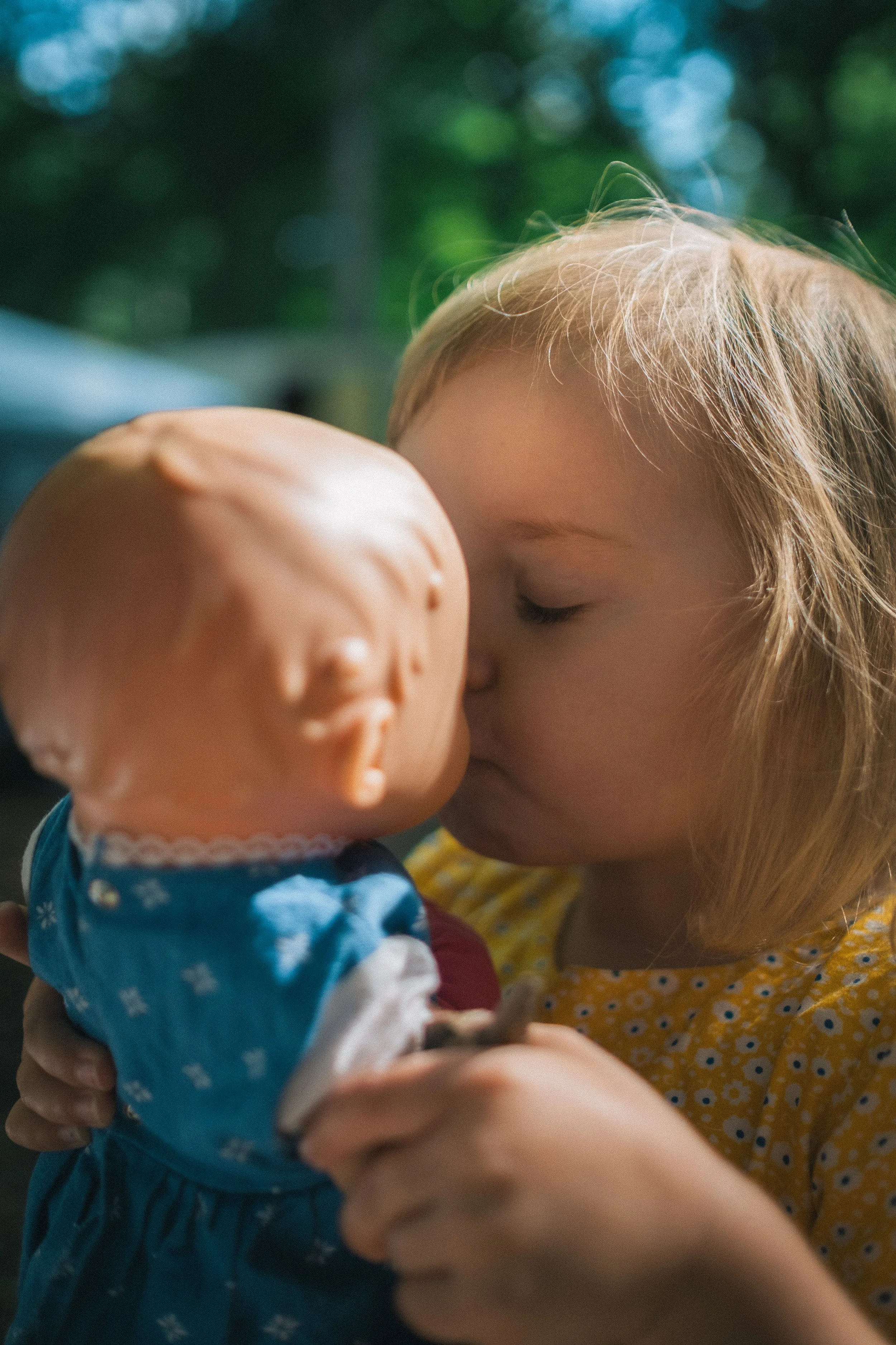 Ein junges Mädchen küsst einen Puppenkopf auf die Lippen, draußen mit unscharfem grünen Hintergrund.