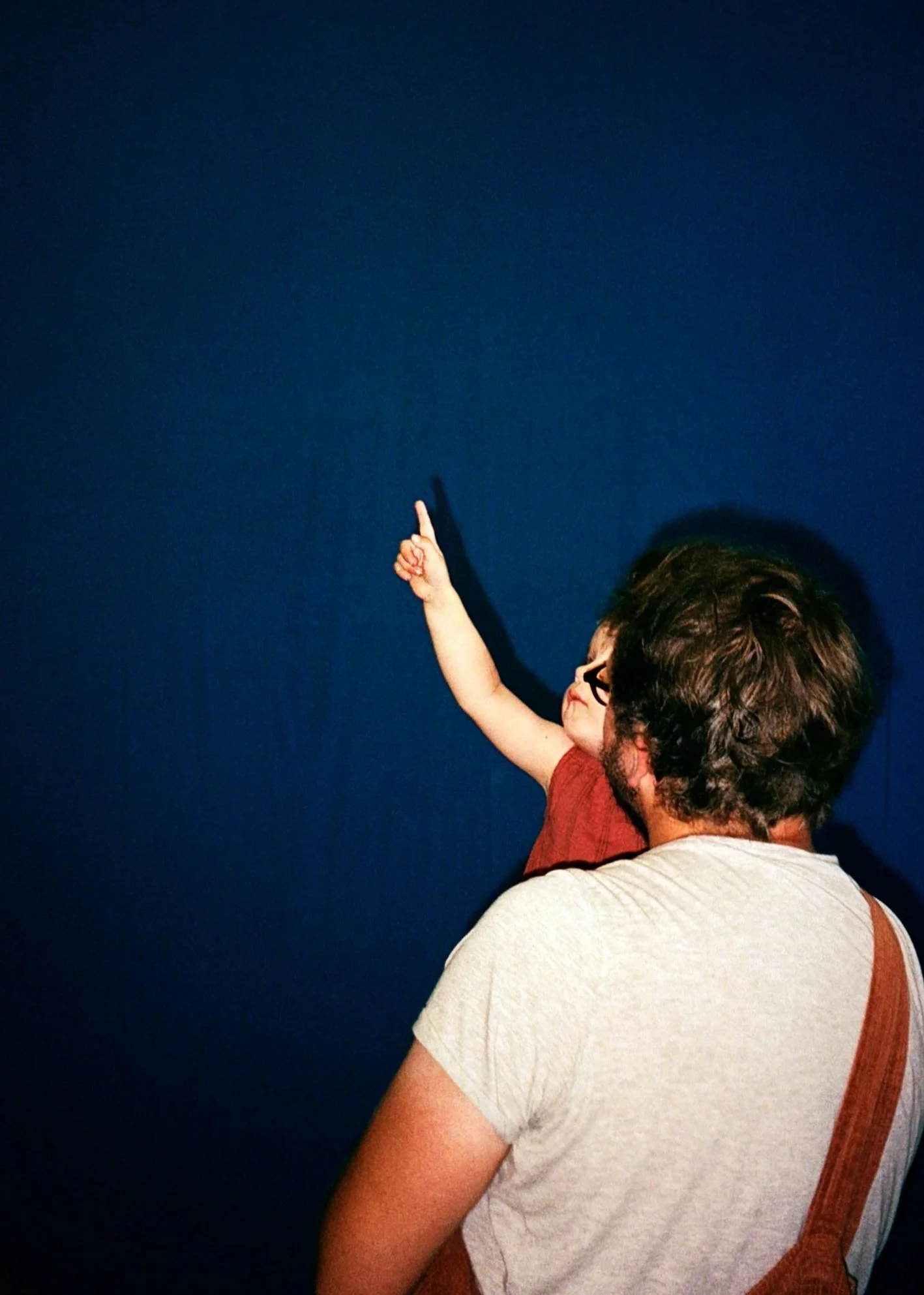 Vor einem dunkelblauem Vorhang steht eiin Mann in weißem T-Shirt. Er trägt ein Kind auf dem Arm welches nach oben zeigt. Die Gesichter sind verdeckt.