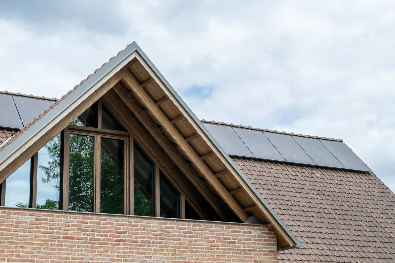 Close-up of a house roof with solar panels, brick wall, and triangular window with sliding glass panels reflecting trees, under cloudy sky.