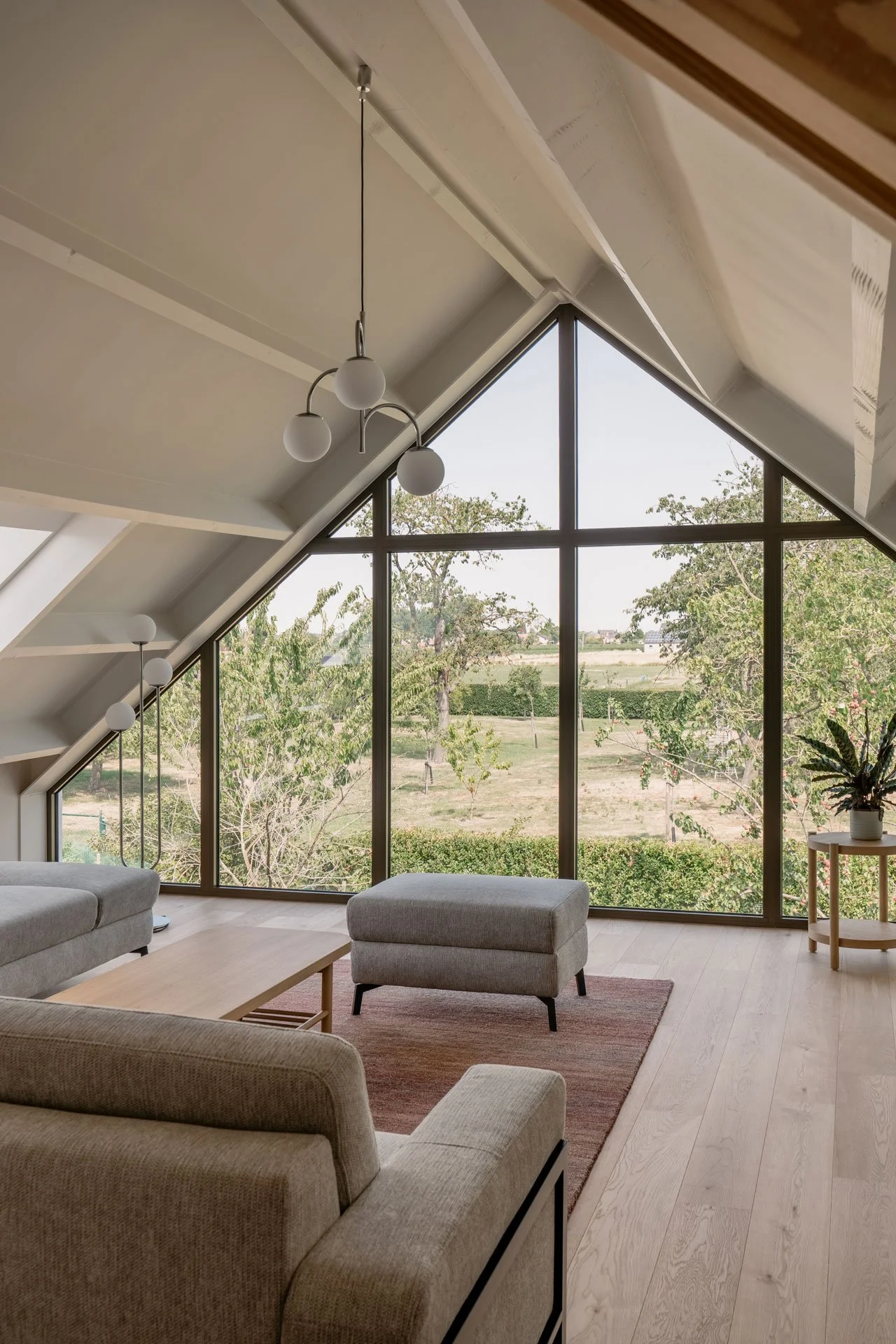 Living room with large triangular window, beige sofas, wooden coffee table, small side table with potted plant, wooden ceiling, and modern hanging light fixtures, overlooking green trees and open landscape.