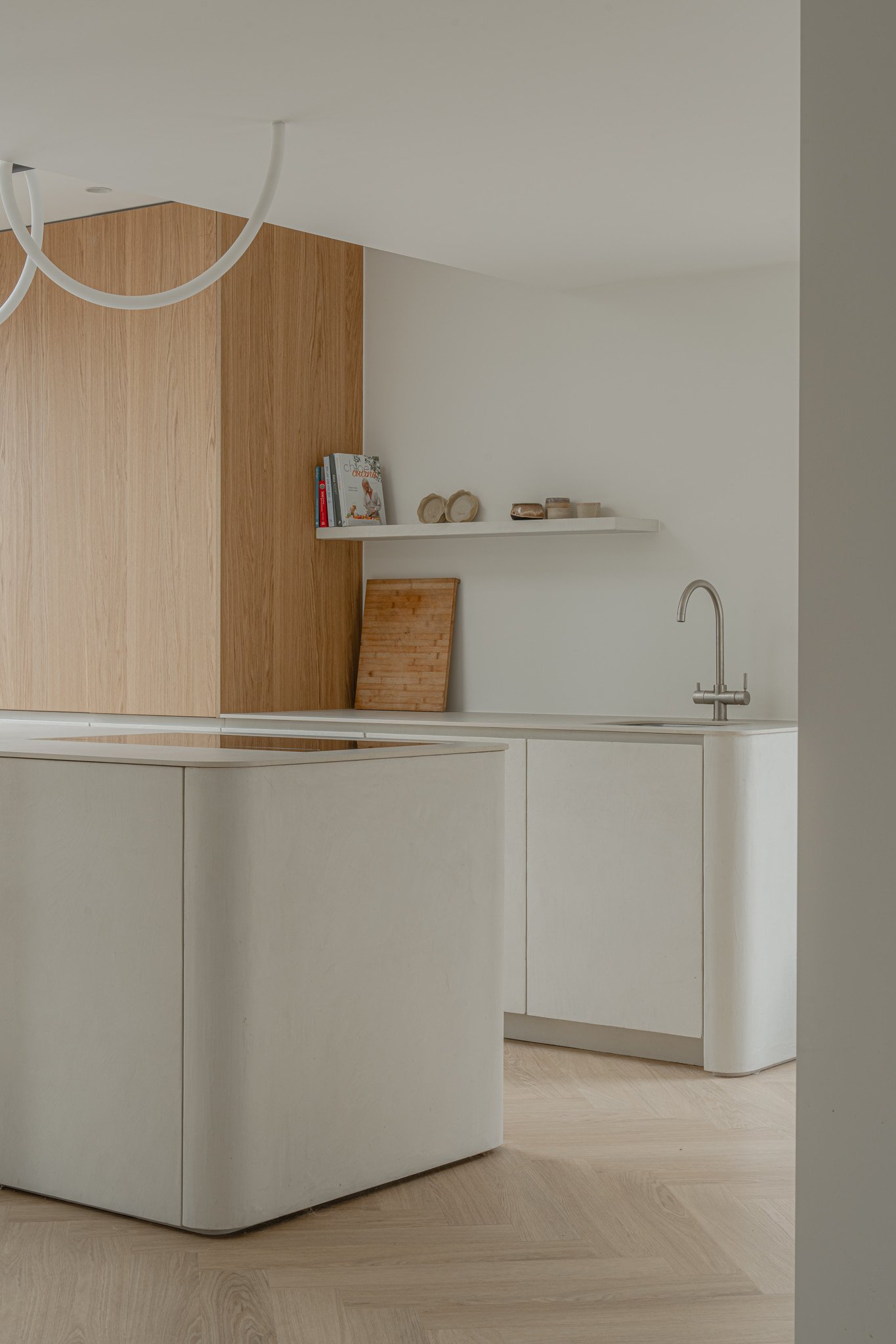 Minimalist kitchen with white cabinets, a white countertop, and a small shelf holding books and decorative items. A wooden panel and cutting board are also visible.