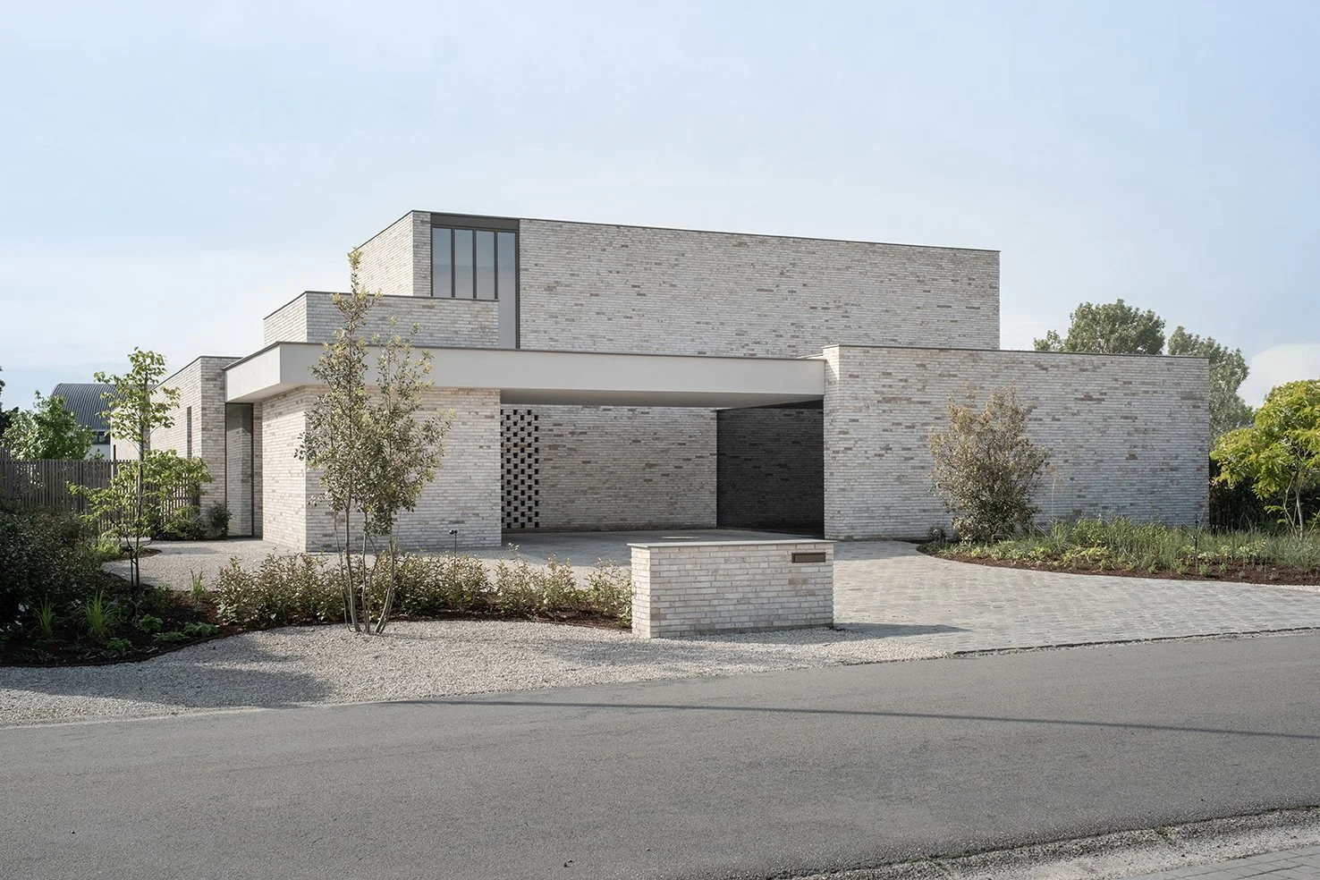 Modern two-story house with light gray brick exterior, flat roof, and minimal landscaping in front, adjacent to a paved road.