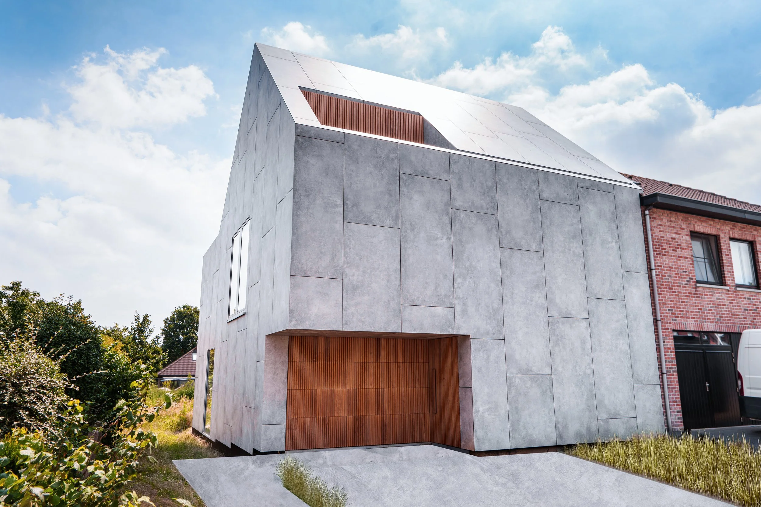 Modern house with gray concrete exterior, large wooden garage door, and sloped roof, situated in a suburban neighborhood with blue sky and clouds.