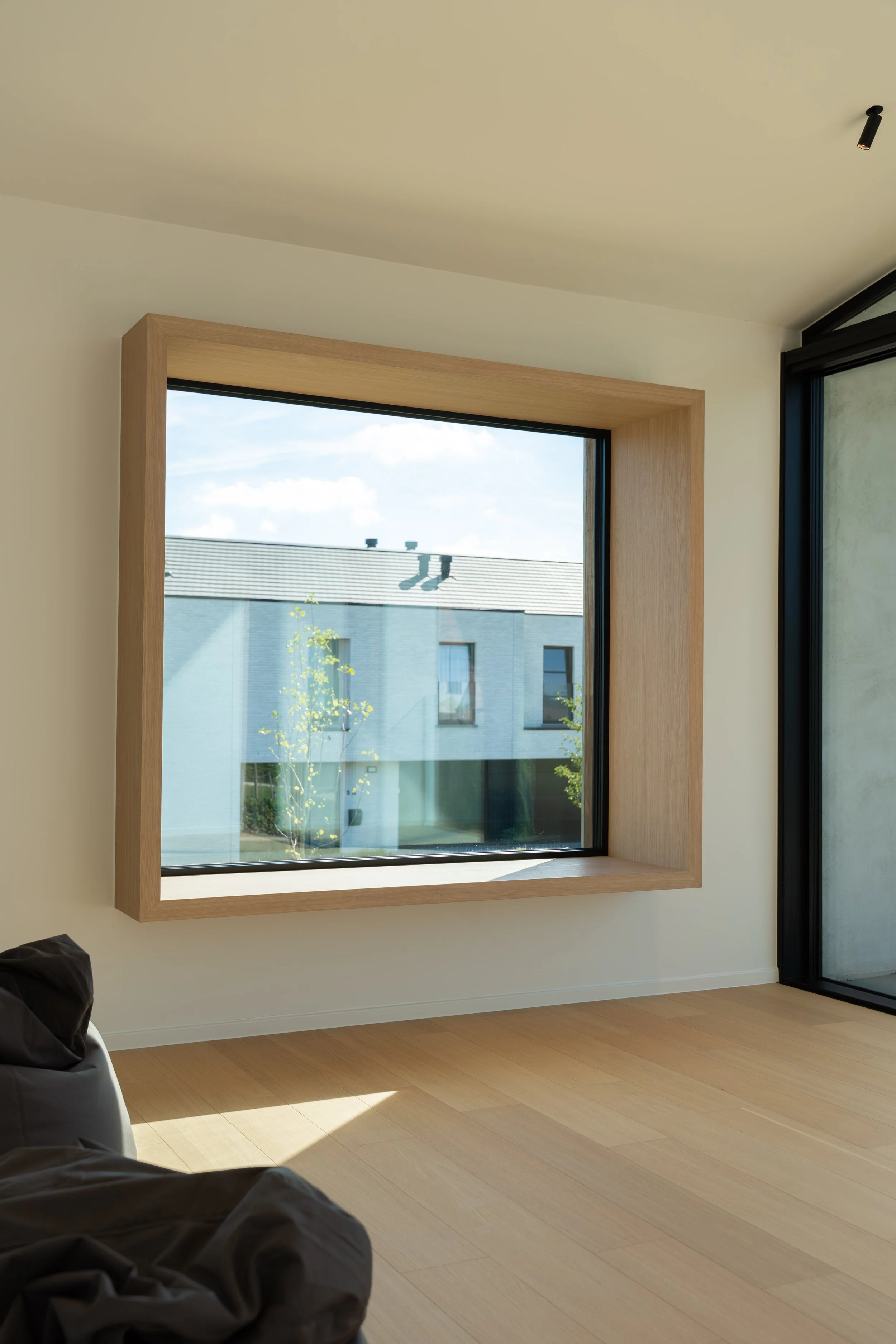 A modern room with light wood flooring, a large square window with a wooden frame, showing a view of a neighboring building and trees outside, and a sliding glass door on the right.