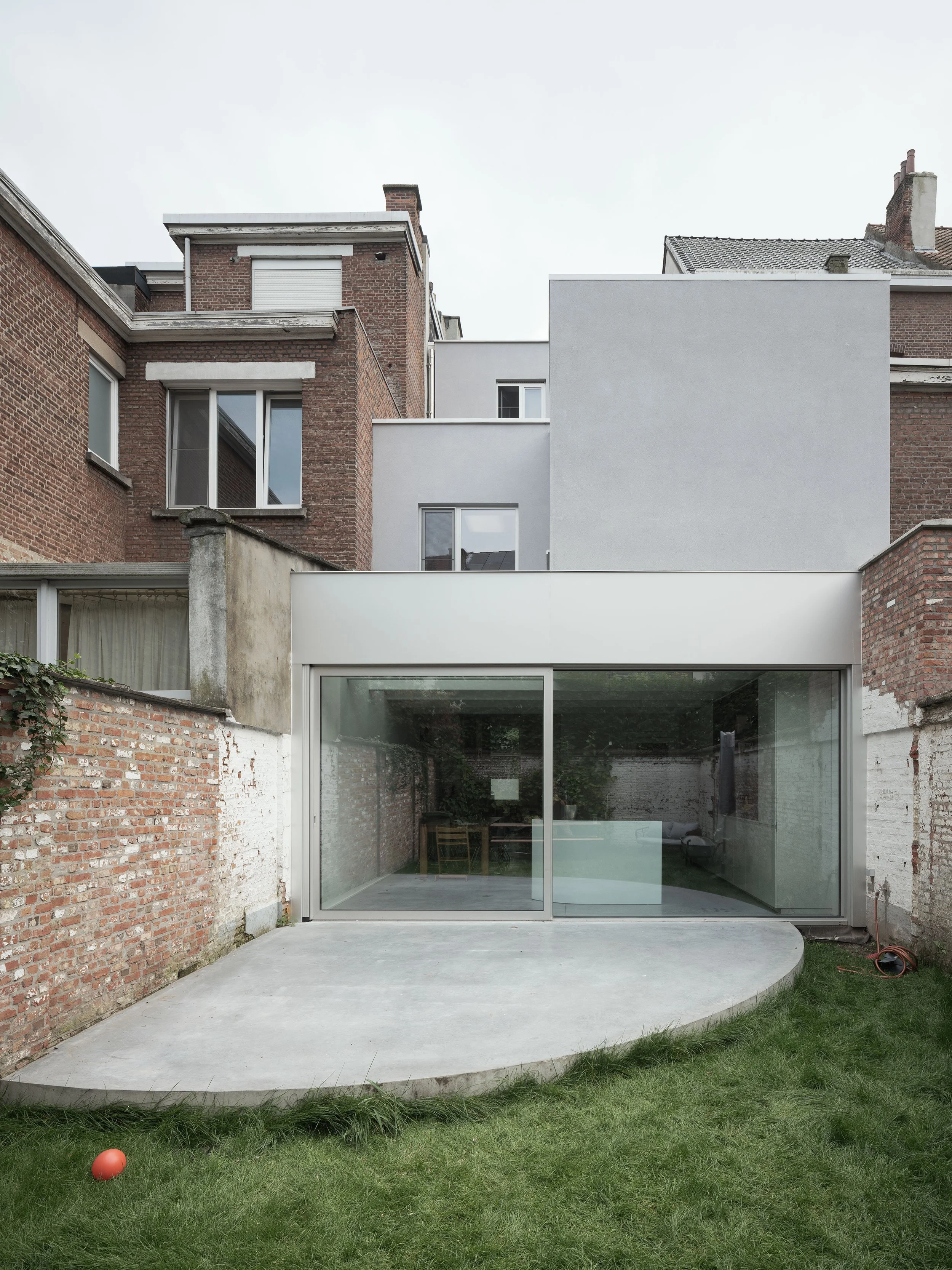 A modern extension with a large glass sliding door on a concrete patio within a backyard, surrounded by brick and concrete walls, with neighboring brick buildings overhead.