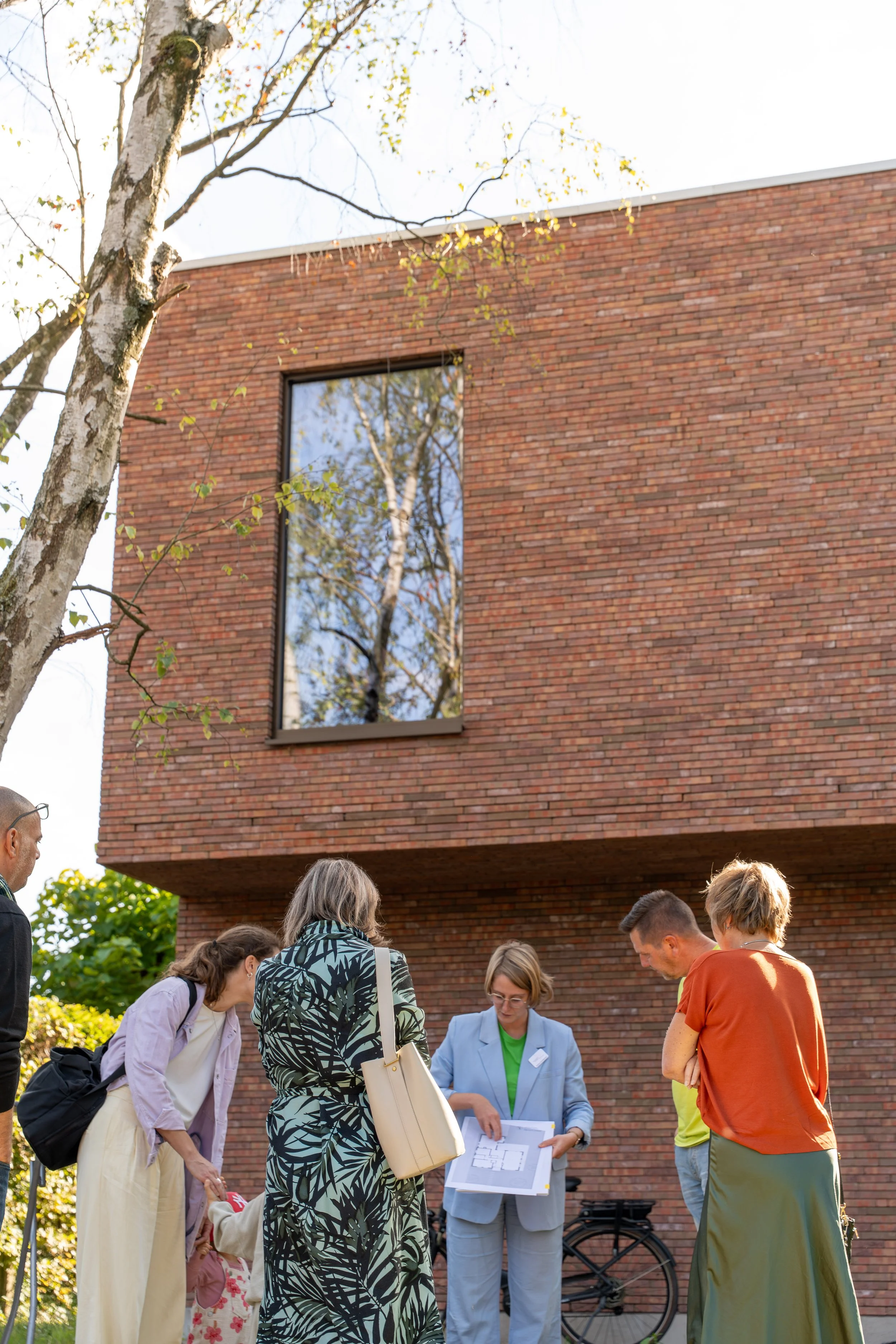 Group of people gathered outside a brick building with large windows, looking at architectural plans during daytime.