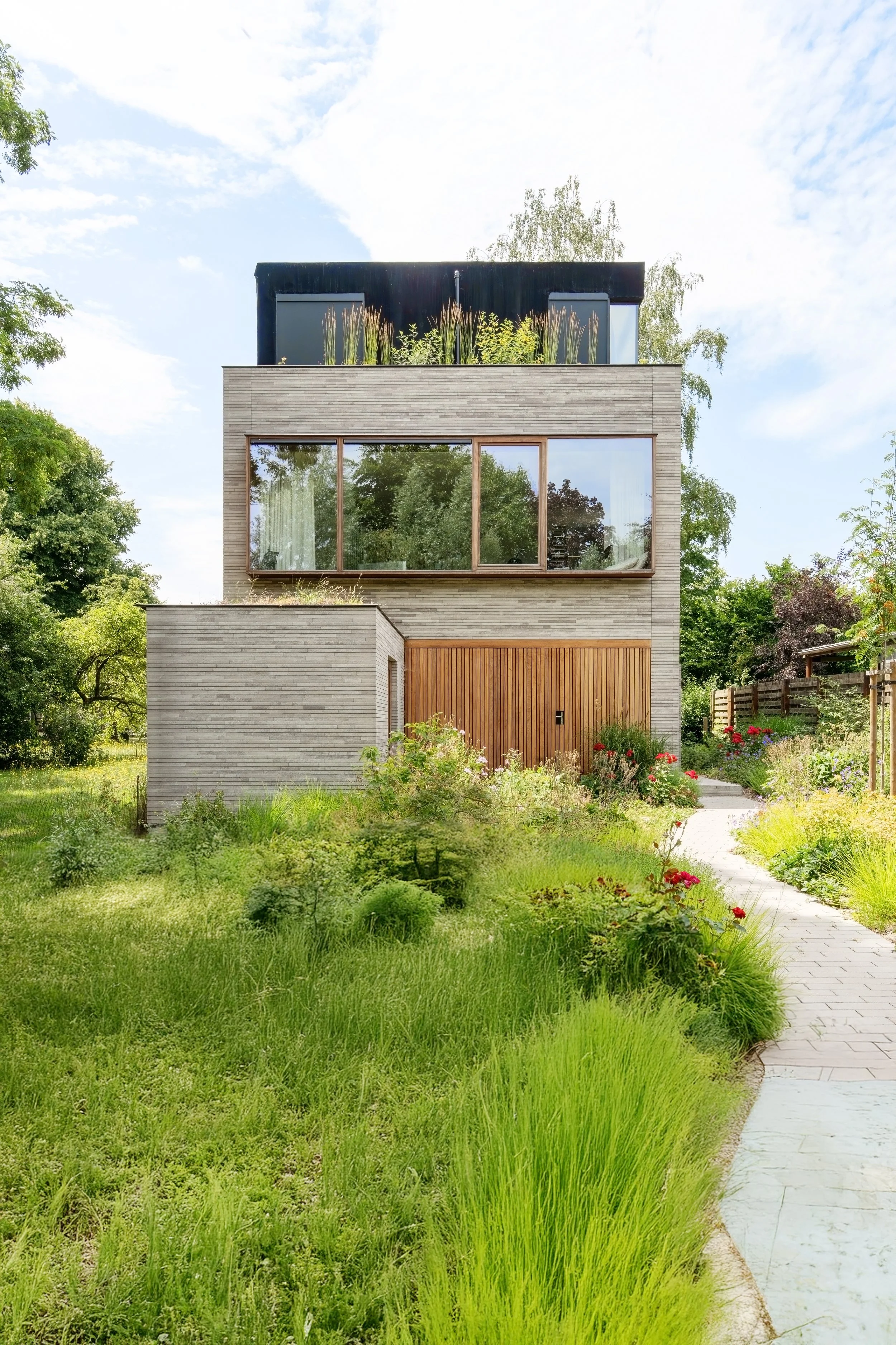 Modern multi-story house with large glass windows, surrounded by a lush green garden and a winding paved pathway.