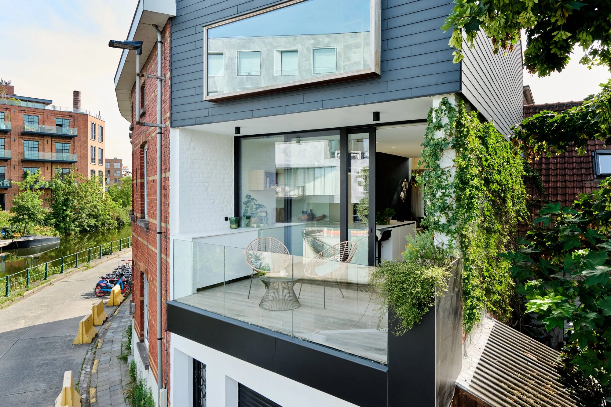 Modern apartment balcony with glass railing, two wicker chairs, and potted plants, overlooking a canal and neighboring buildings.