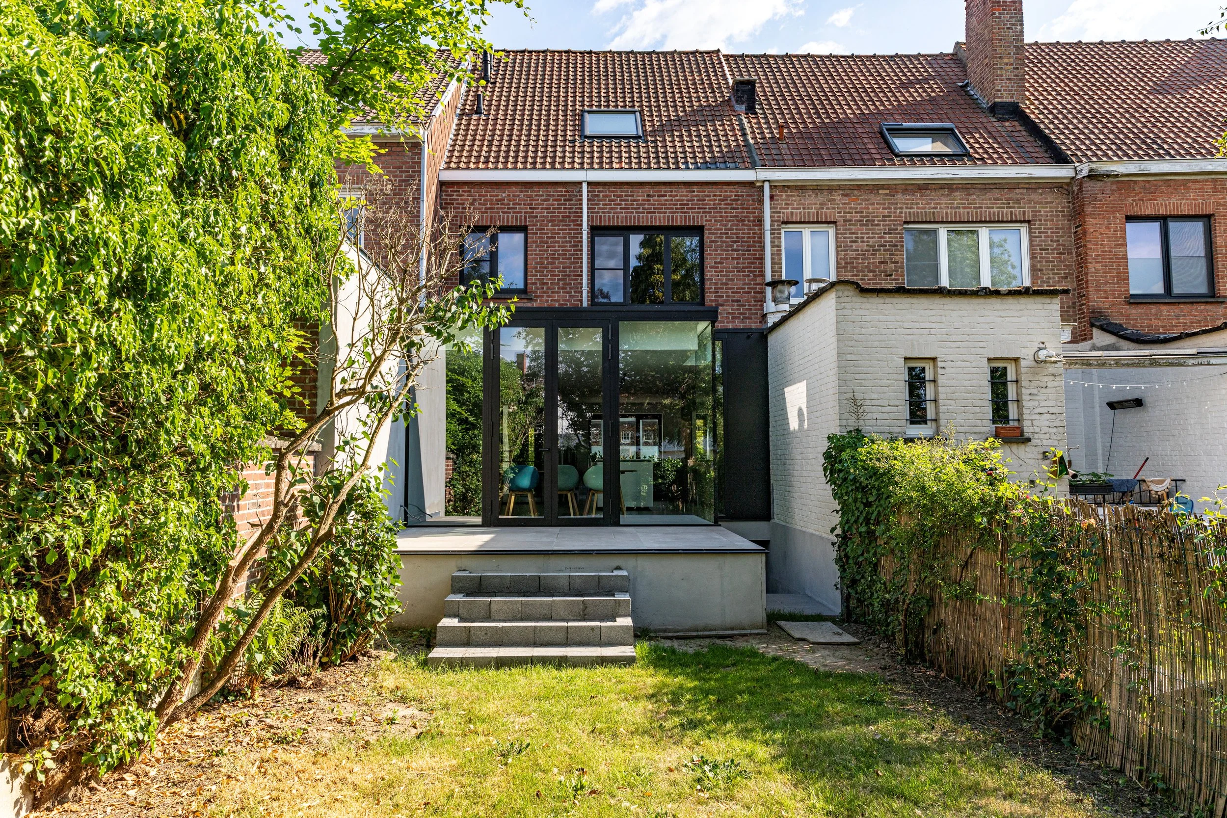 Backyard with steps leading to a glass-enclosed patio area attached to a brick house. The yard has grass, bushes, and a tree on the left side, with neighboring houses visible in the background.