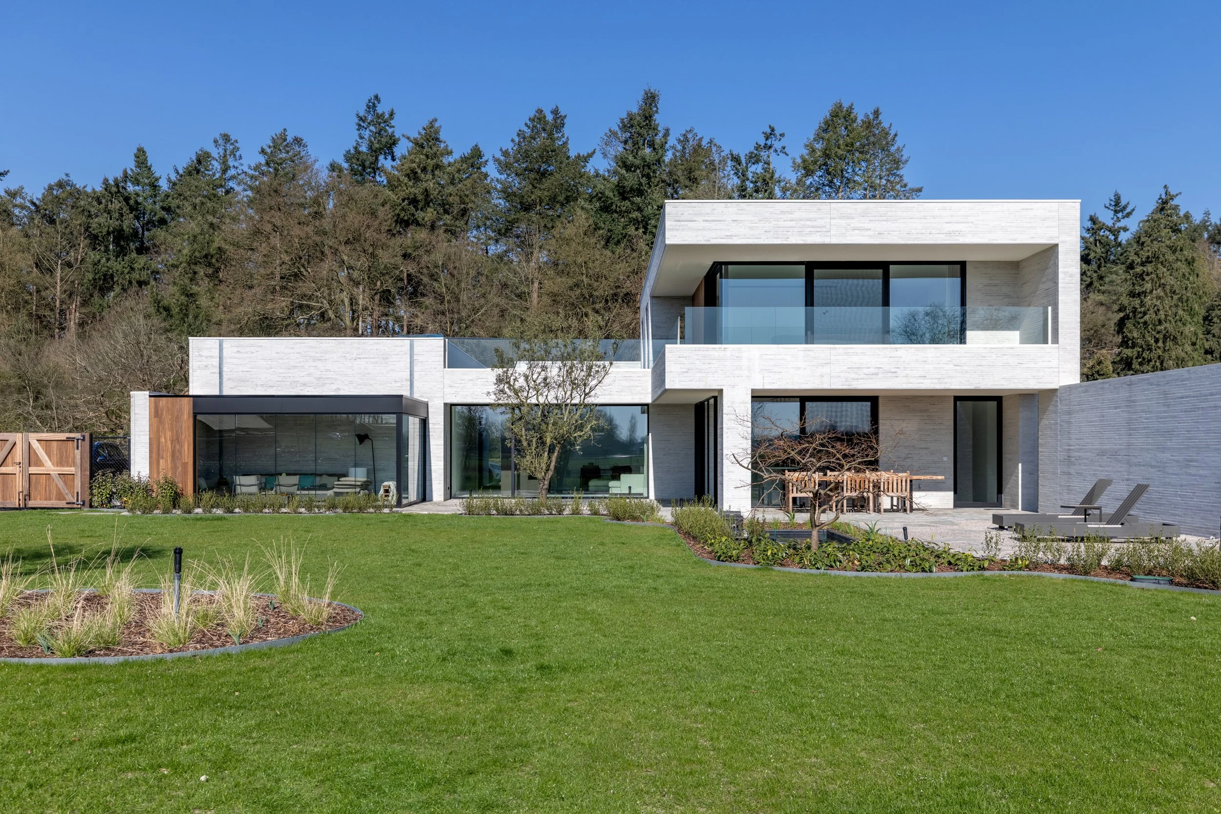 Modern white house with large windows and a landscaped yard with flowers and green grass, with trees and a clear blue sky in the background.