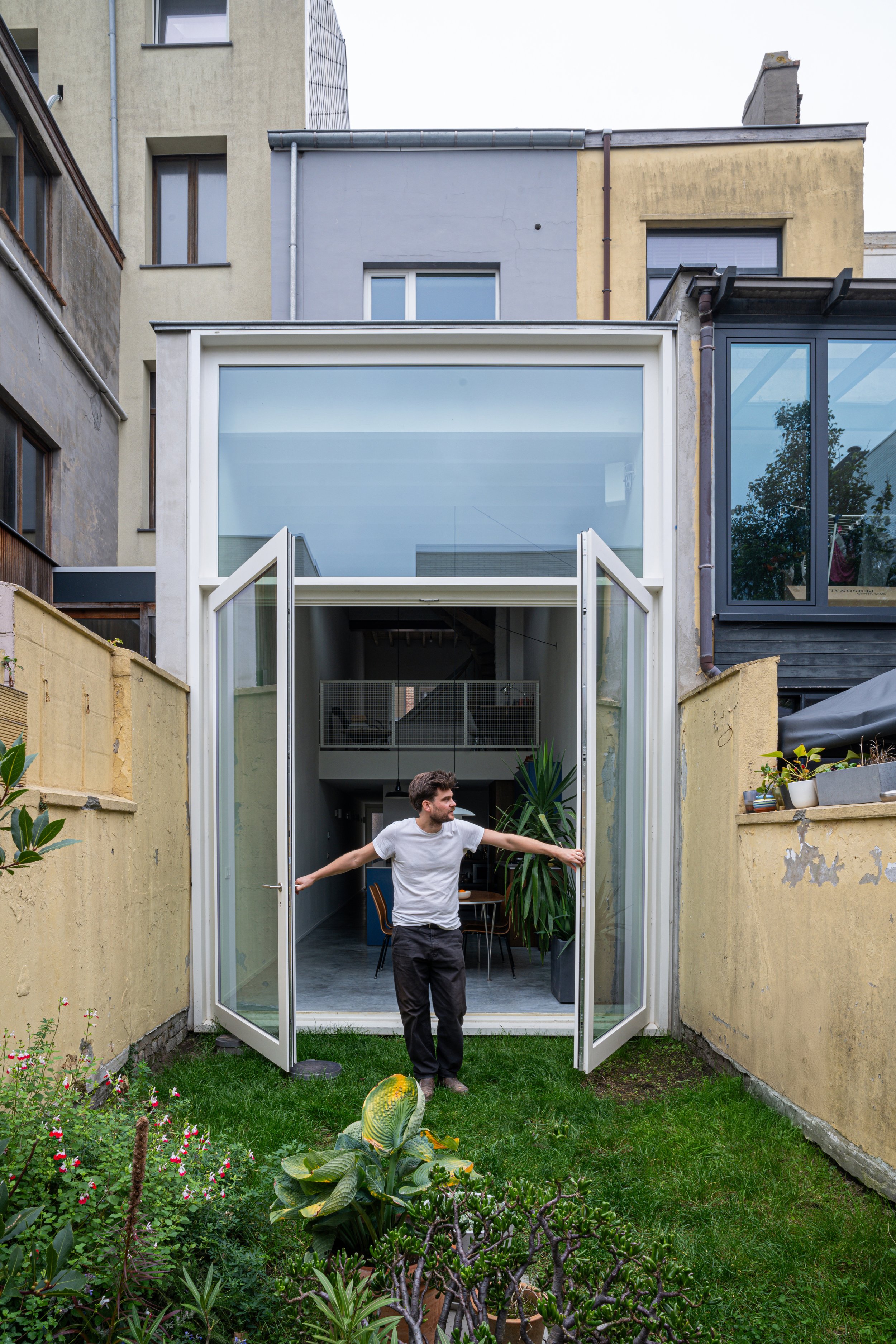 A man opening glass doors in a small backyard garden, with a modern house in the background.