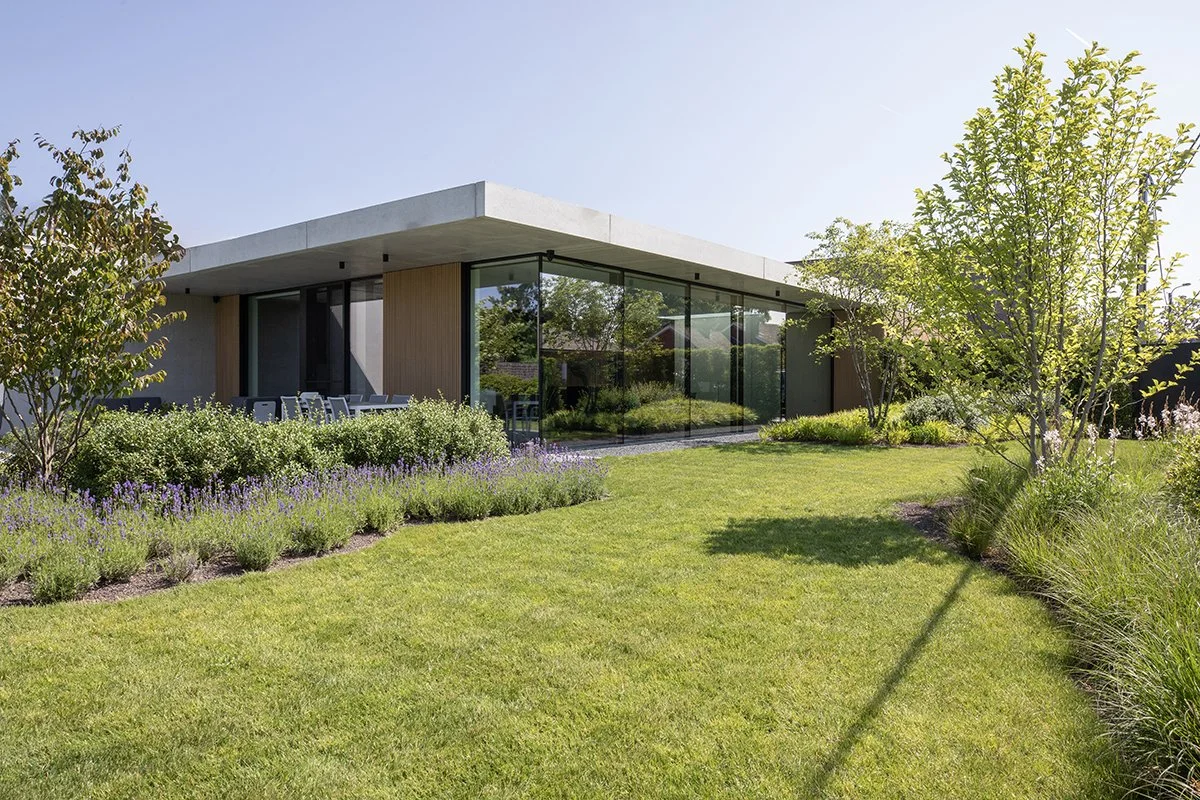 Modern house with glass walls, surrounded by a well-maintained garden with green grass, trees, and shrubs, under a clear blue sky.